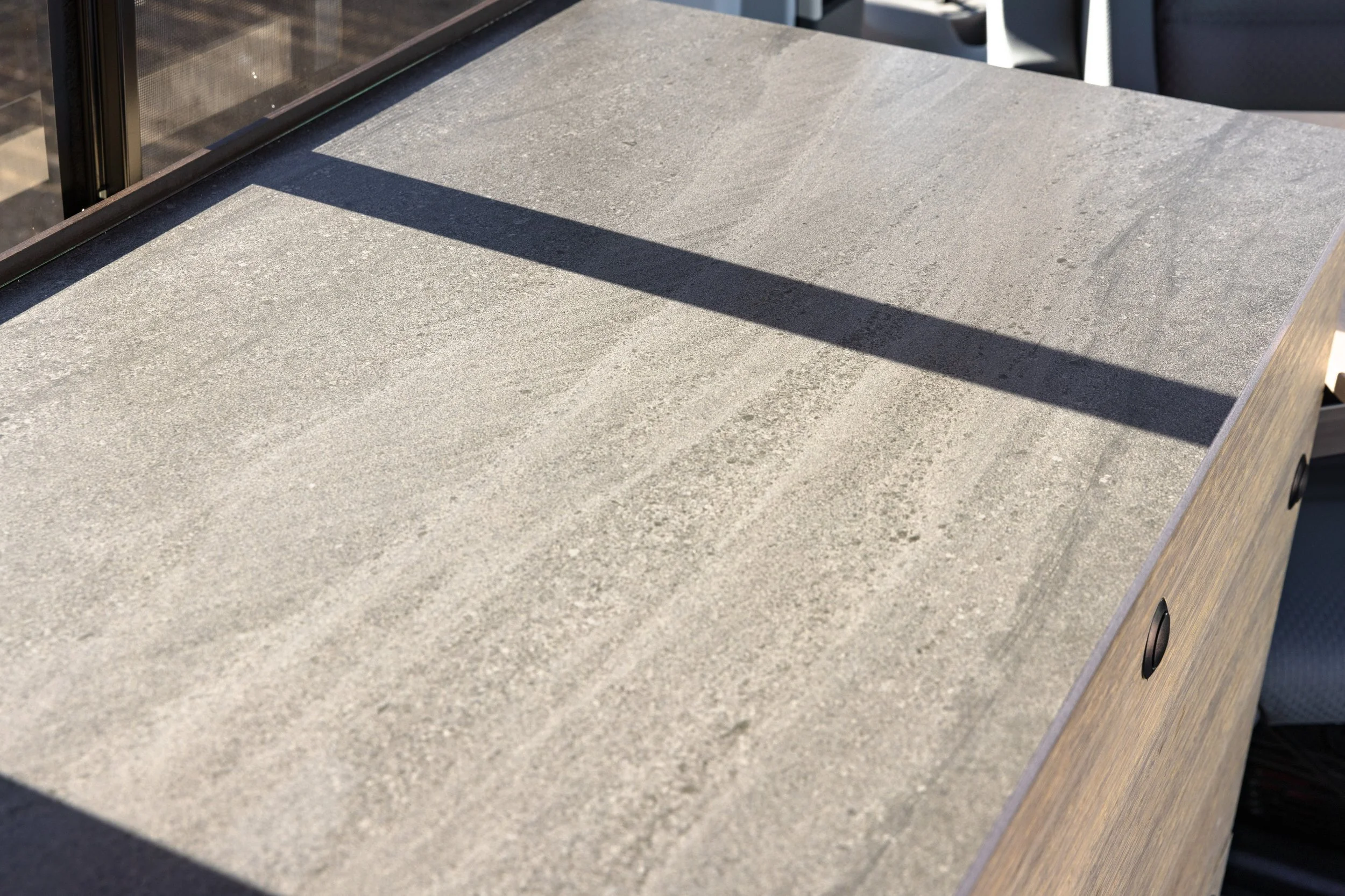 A close-up view of a gray stone countertop with a shadow cast across it. Part of a glass wall and a chair are visible at the edges.
