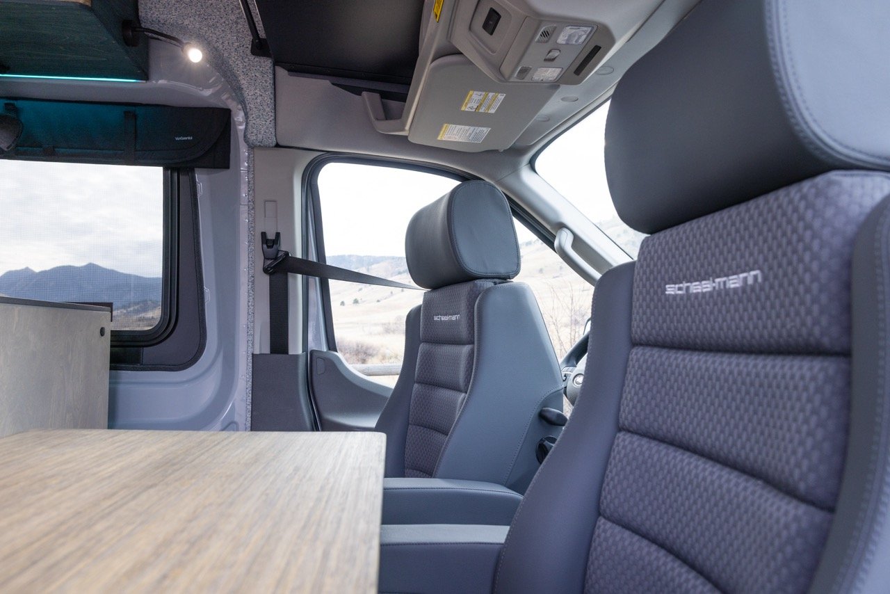 Interior of a camper van with two gray seats and a wooden table, large window showing a mountain landscape outside.