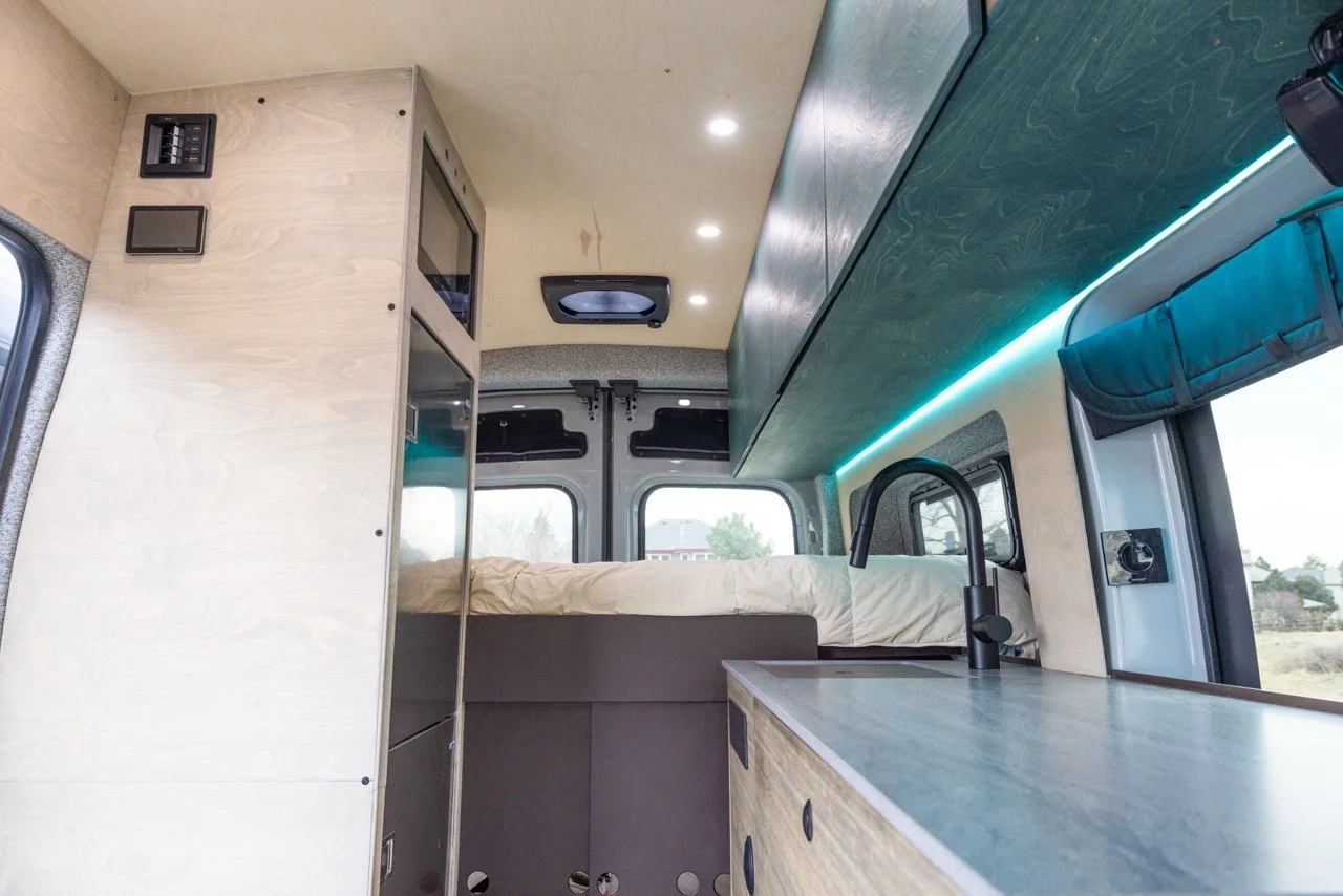 Interior of a camper van with a bed, a kitchen sink, overhead storage, and windows showing outdoor scenery.