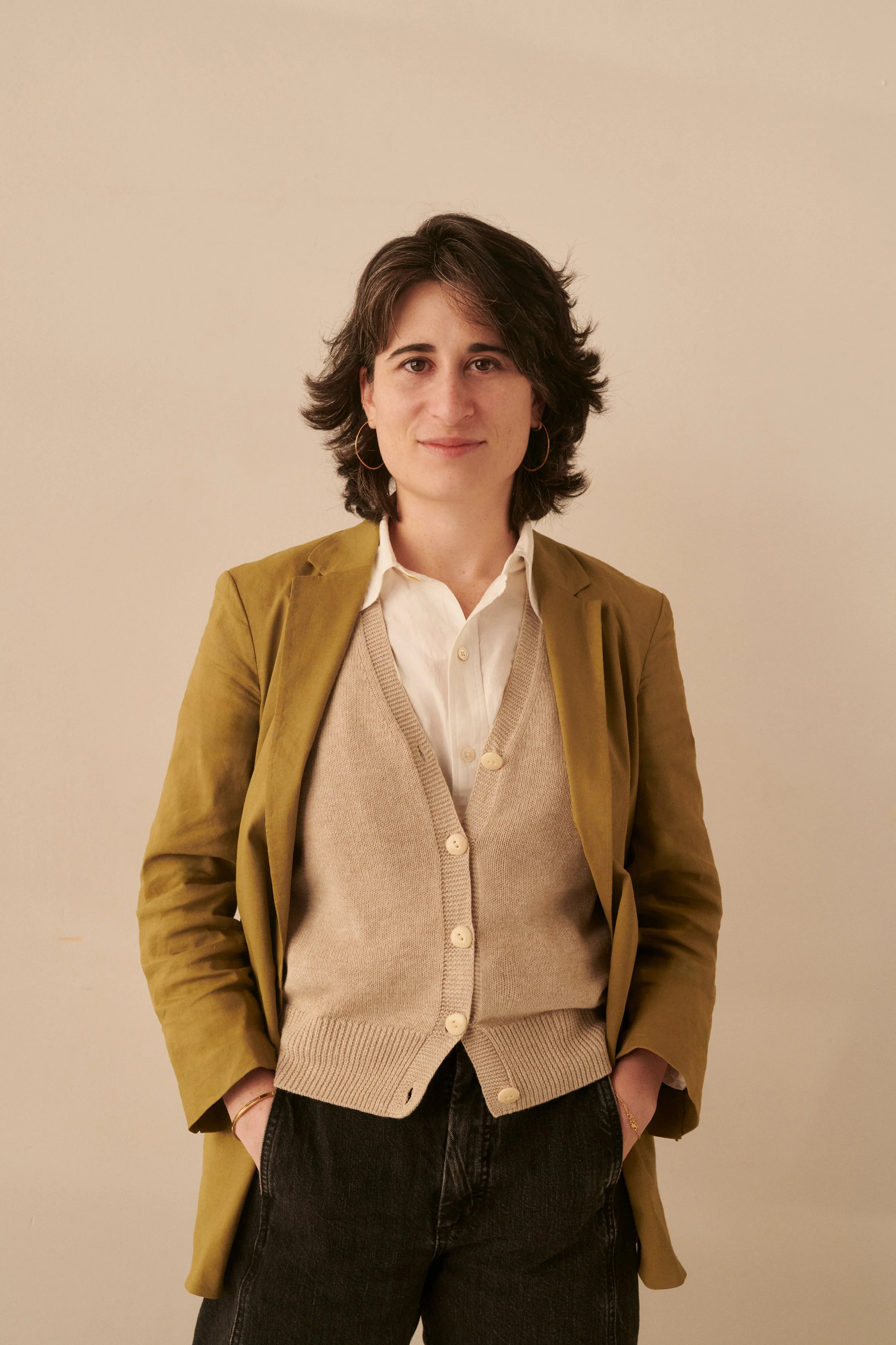 A young woman with dark, wavy hair and hoop earrings, wearing a beige cardigan over a white shirt and yellow blazer, standing against a plain beige wall.