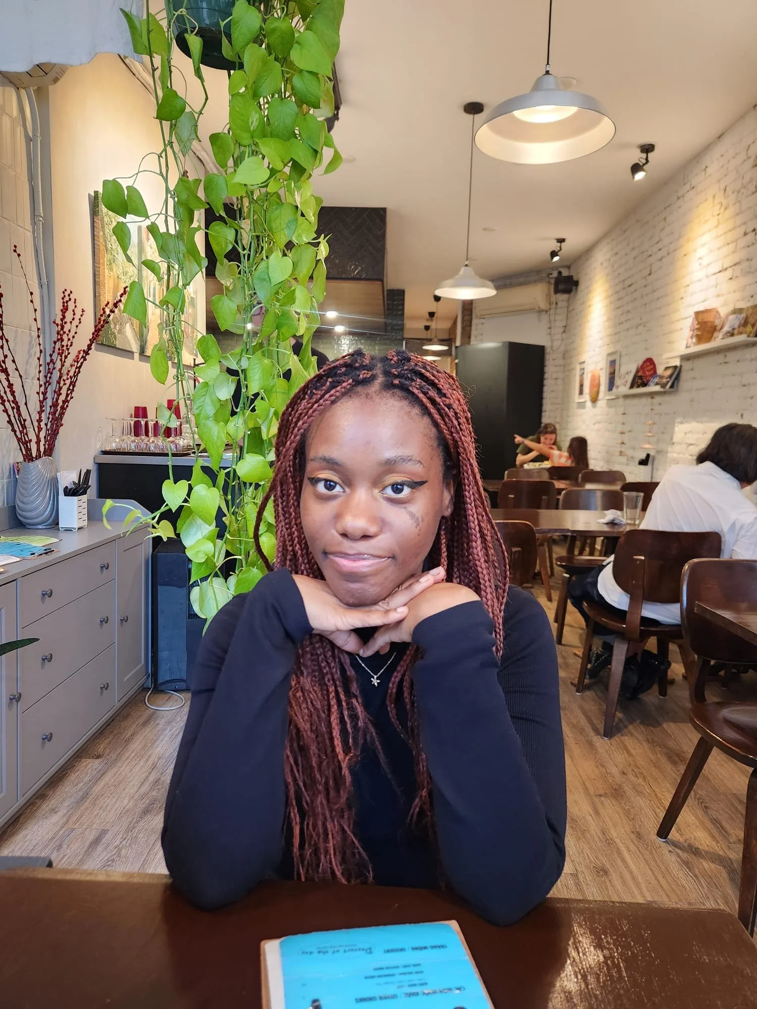 A young femme-presenting person with reddish-brown braids sitting at a table in a cozy cafe, resting their chin on their hands, with plants and artwork in the background.