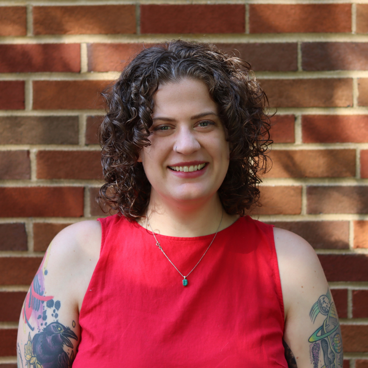 Smiling woman with curly hair wearing a pink blouse standing in front of a brick wall.