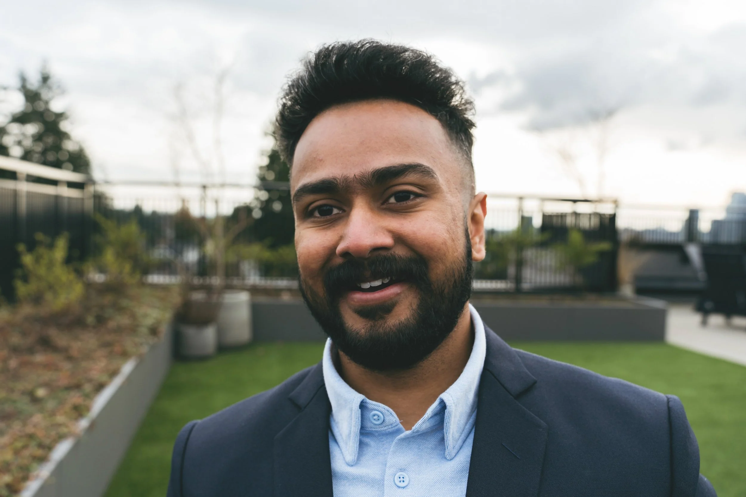 Close-up of a smiling man with dark hair and beard, dressed in a suit, standing outdoors on a cloudy day with trees and a fence in the background.