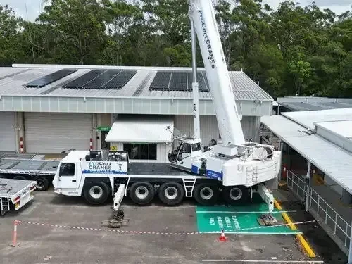 A large crane truck is parked next to a building with solar panels on the roof, using its boom to lift or work on the roof area.