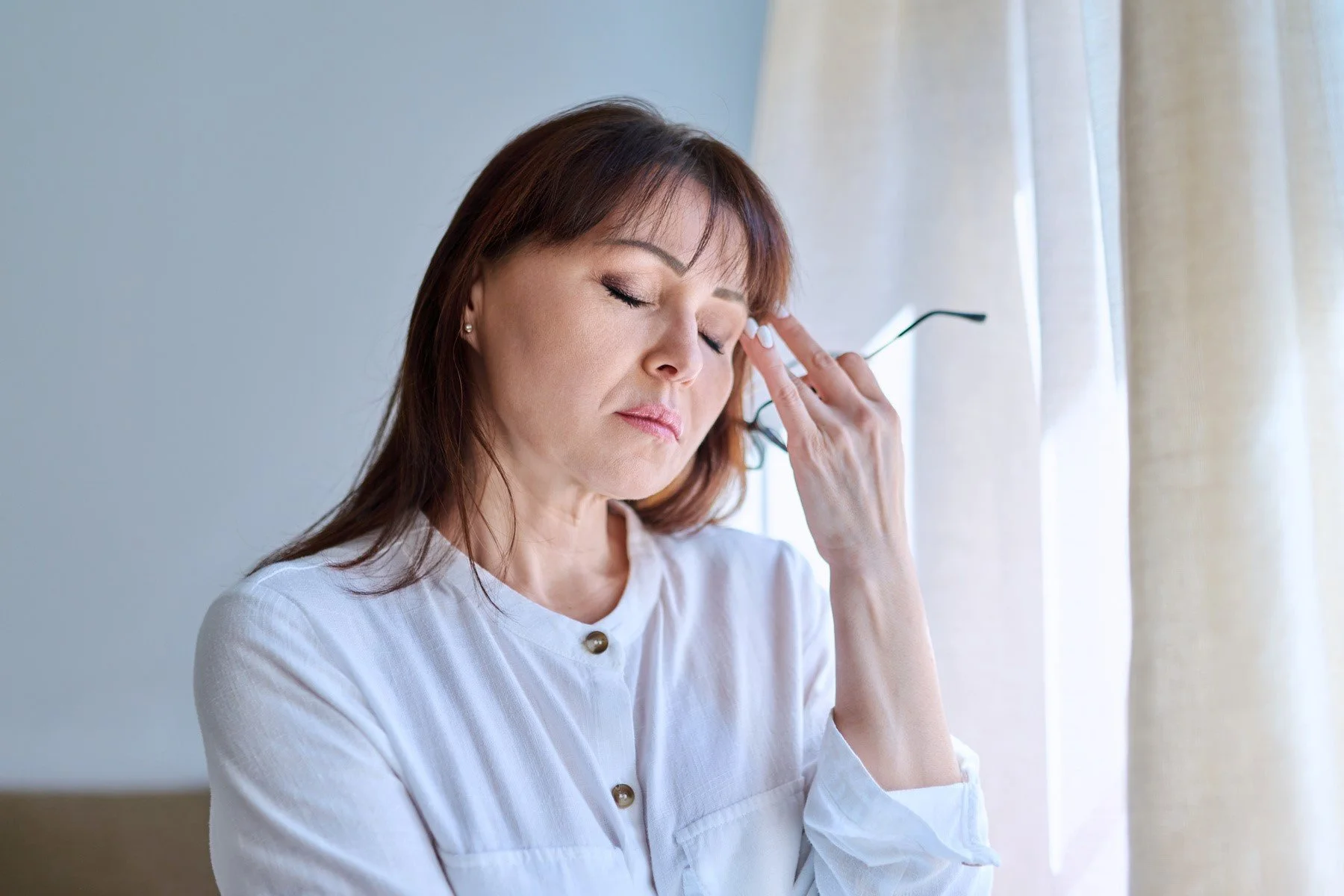 A dark haired woman holding her hand to her brow looking tired with her eyes closed