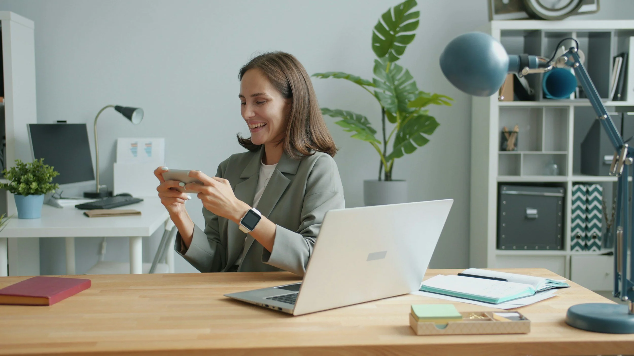 A woman sitting at a work desk, smiling and looking at her phone, with a laptop, notebooks, and a plant in a modern office setting.