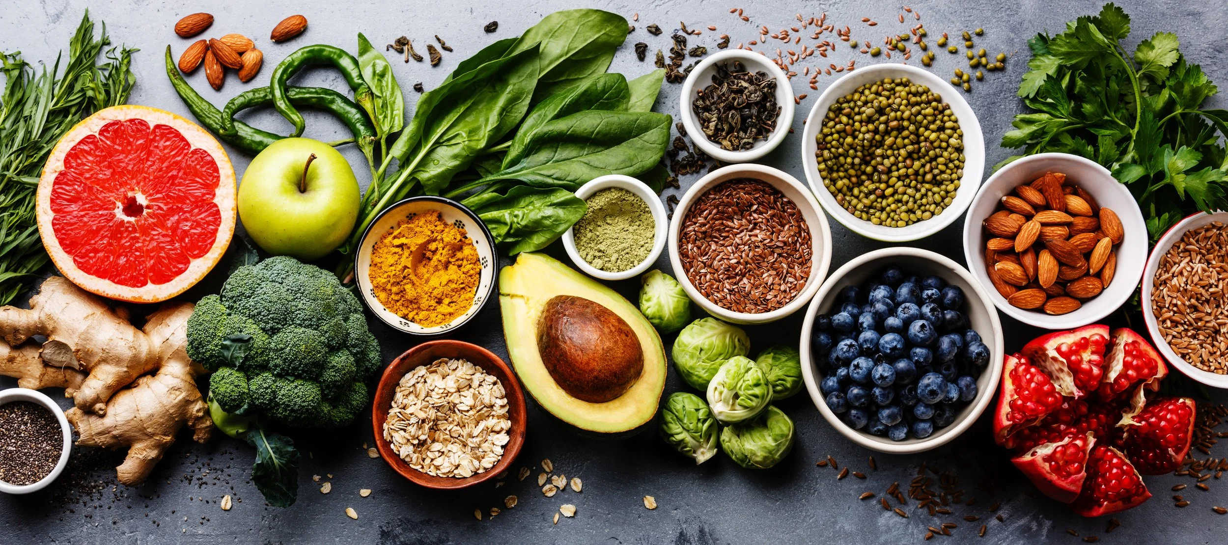 A variety of fresh fruits, vegetables, and seeds spread out on a gray surface, including grapefruit, green apple, avocado, blueberries, pomegranate, ginger, broccoli, spinach, turmeric, alfalfa sprouts, flax seeds, chia seeds, mung beans, lentils, hemp seeds, and other herbs and spices.