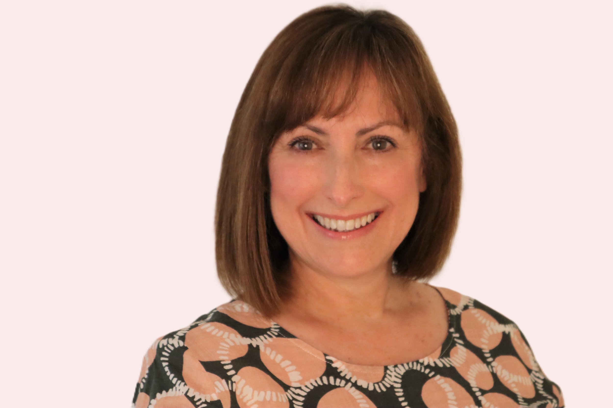A woman with shoulder-length light brown hair and light skin, smiling and wearing a patterned top with black, light pink, and white colors, against a plain light background.