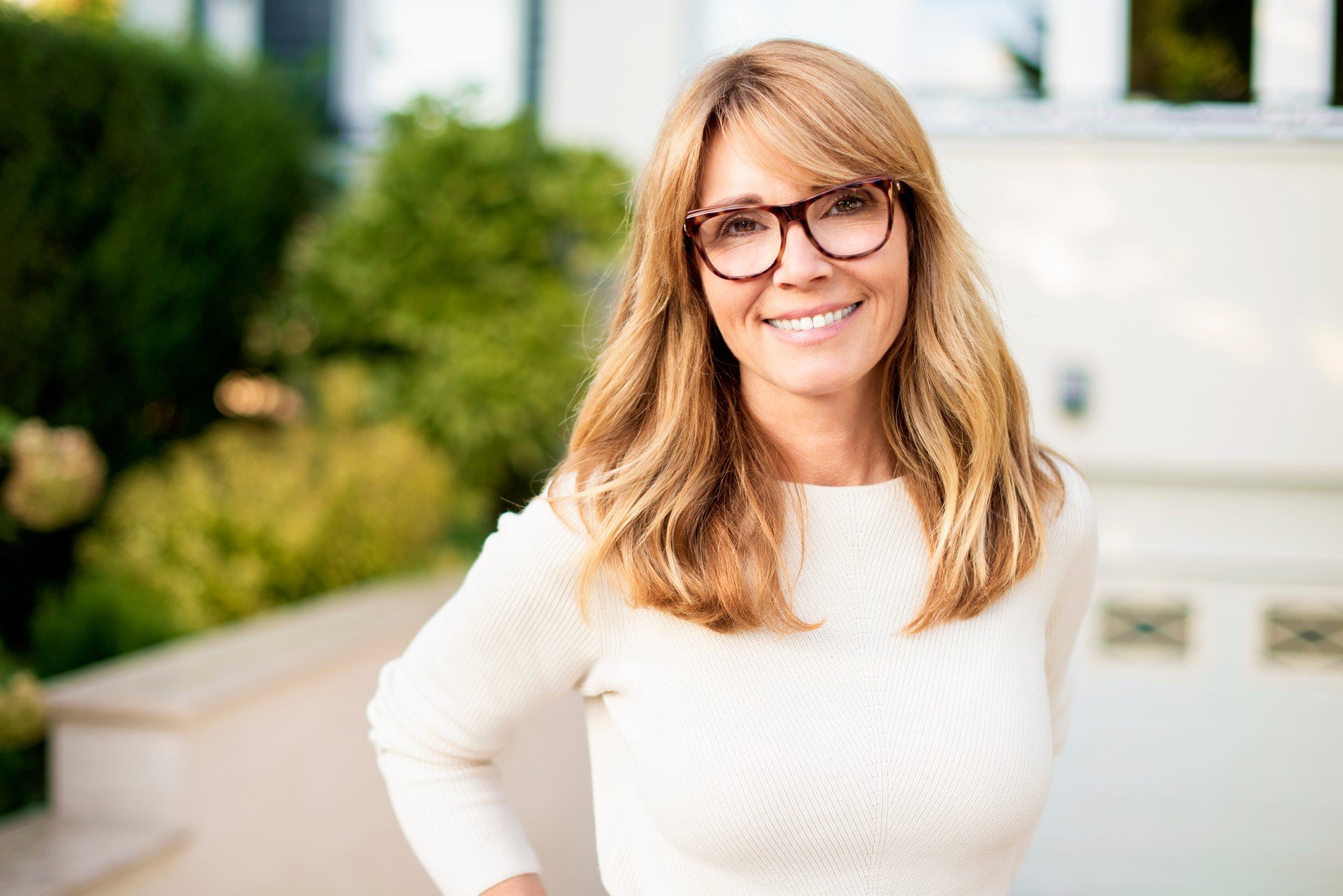 A woman with red hair wearing glasses and a white sweater, smiling outdoors in front of greenery and a white building.