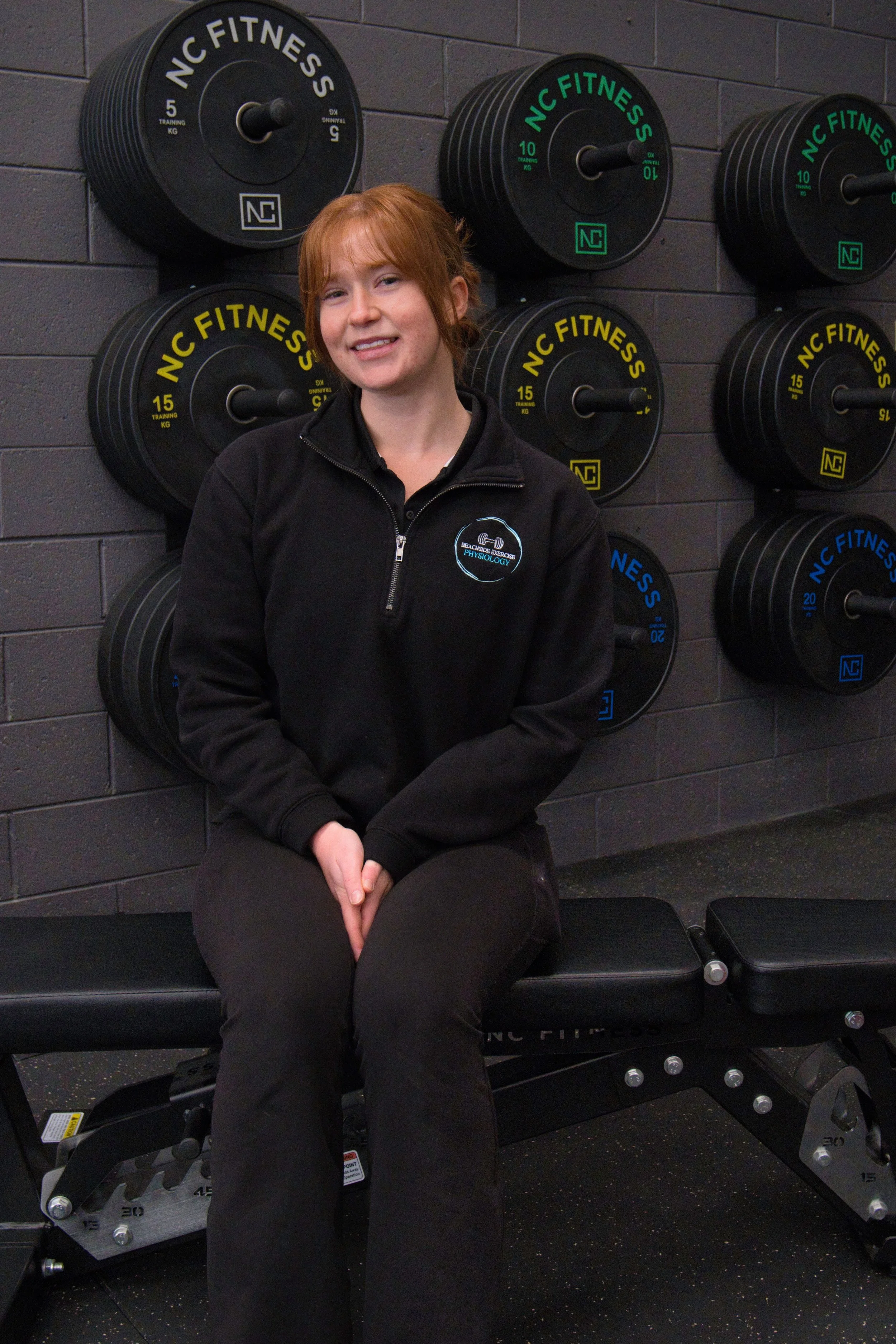 A young woman with red hair sitting on a black workout bench in a gym. She is wearing a black zip-up jacket with the logo 'Beachside Exercise Physiology' and black pants. Meg Benger AES