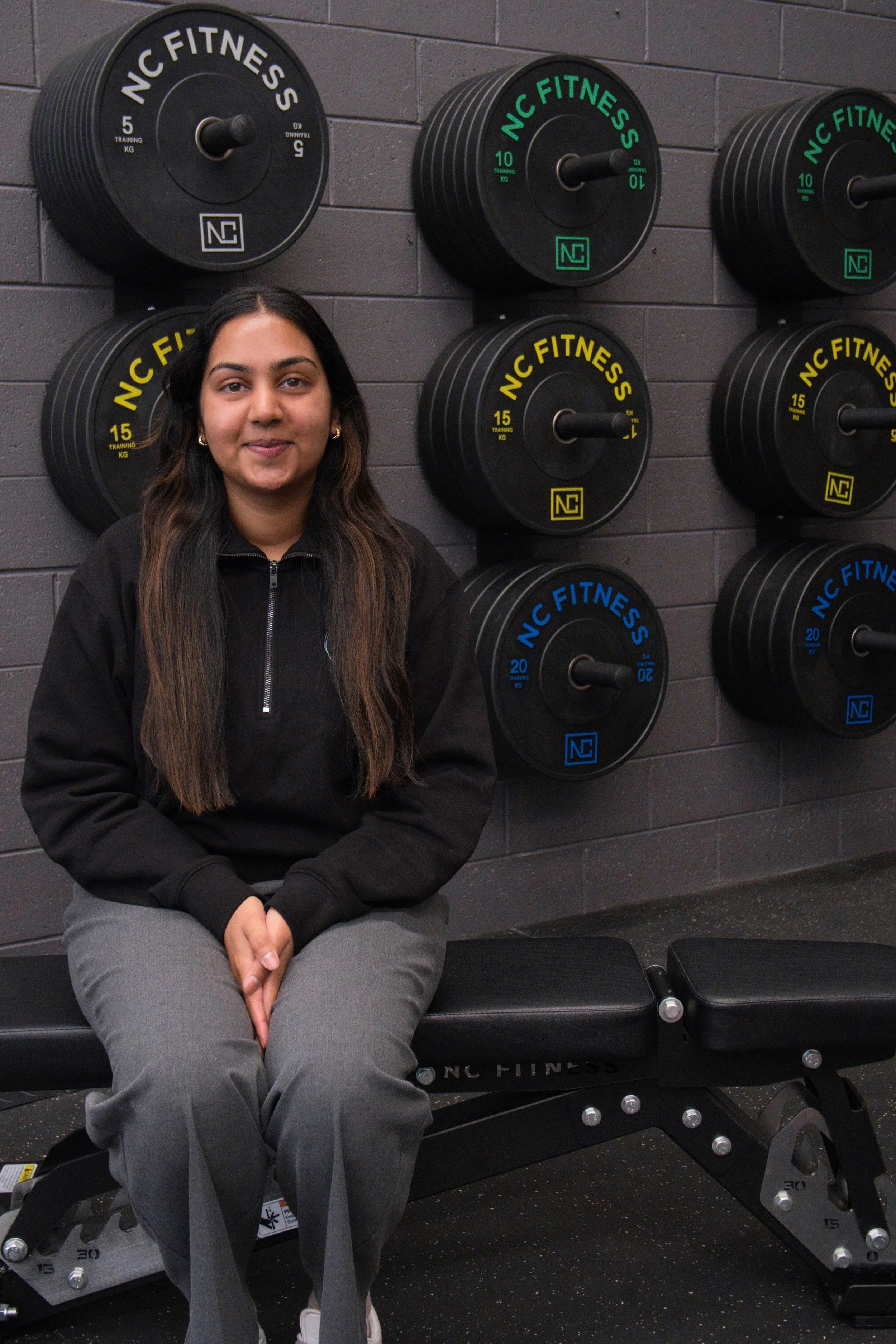 An Accredited Practising Dietitian sitting on a black workout bench at a fitness center, with weights on the wall behind her labelled 'NC FITNESS' in various colors. - Samadi Mallawa APD.