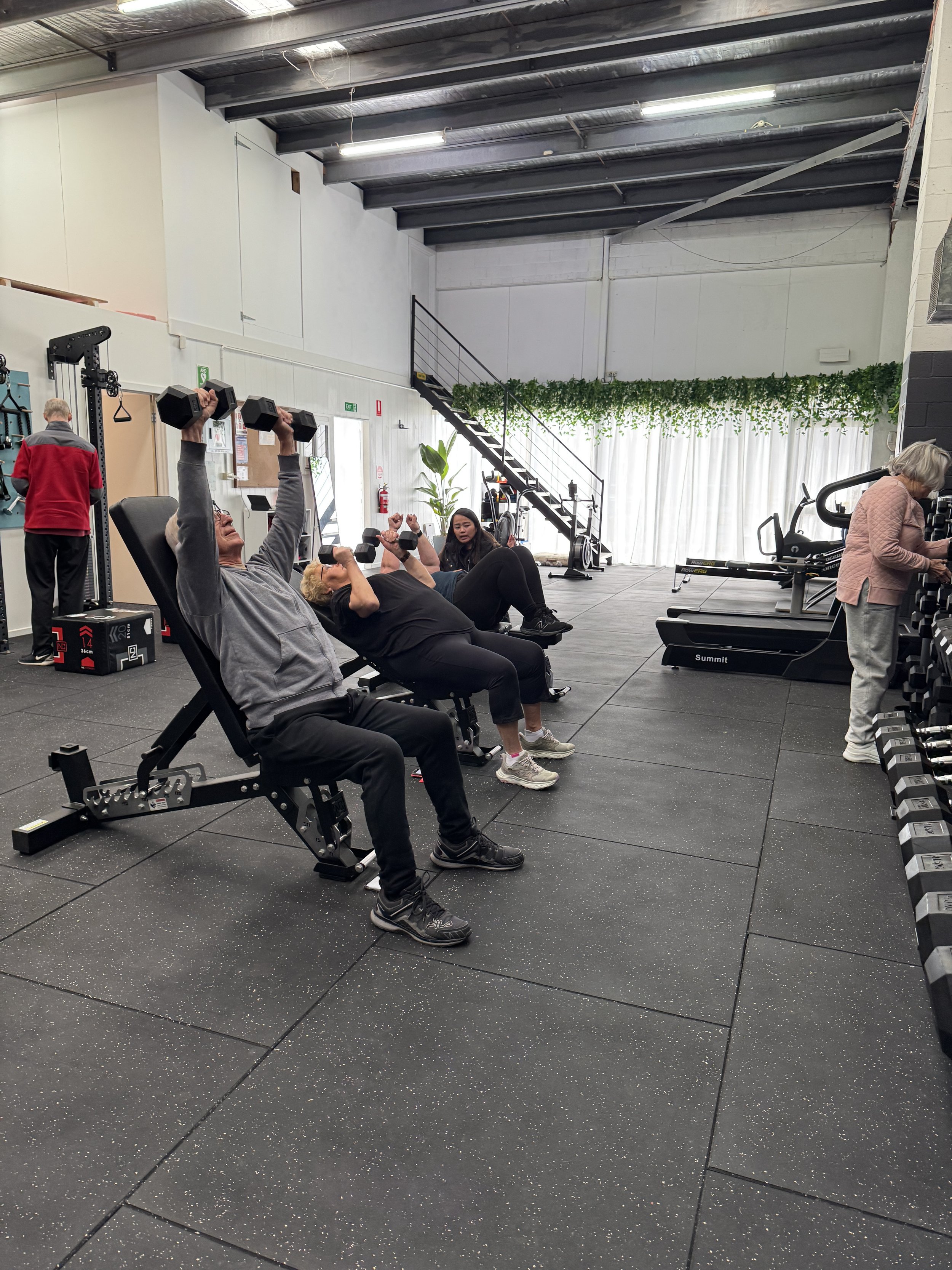 People working out with exercise equipment at a gym, including a man holding a dumbbell in a squat position and an exercise physiologist observing him.