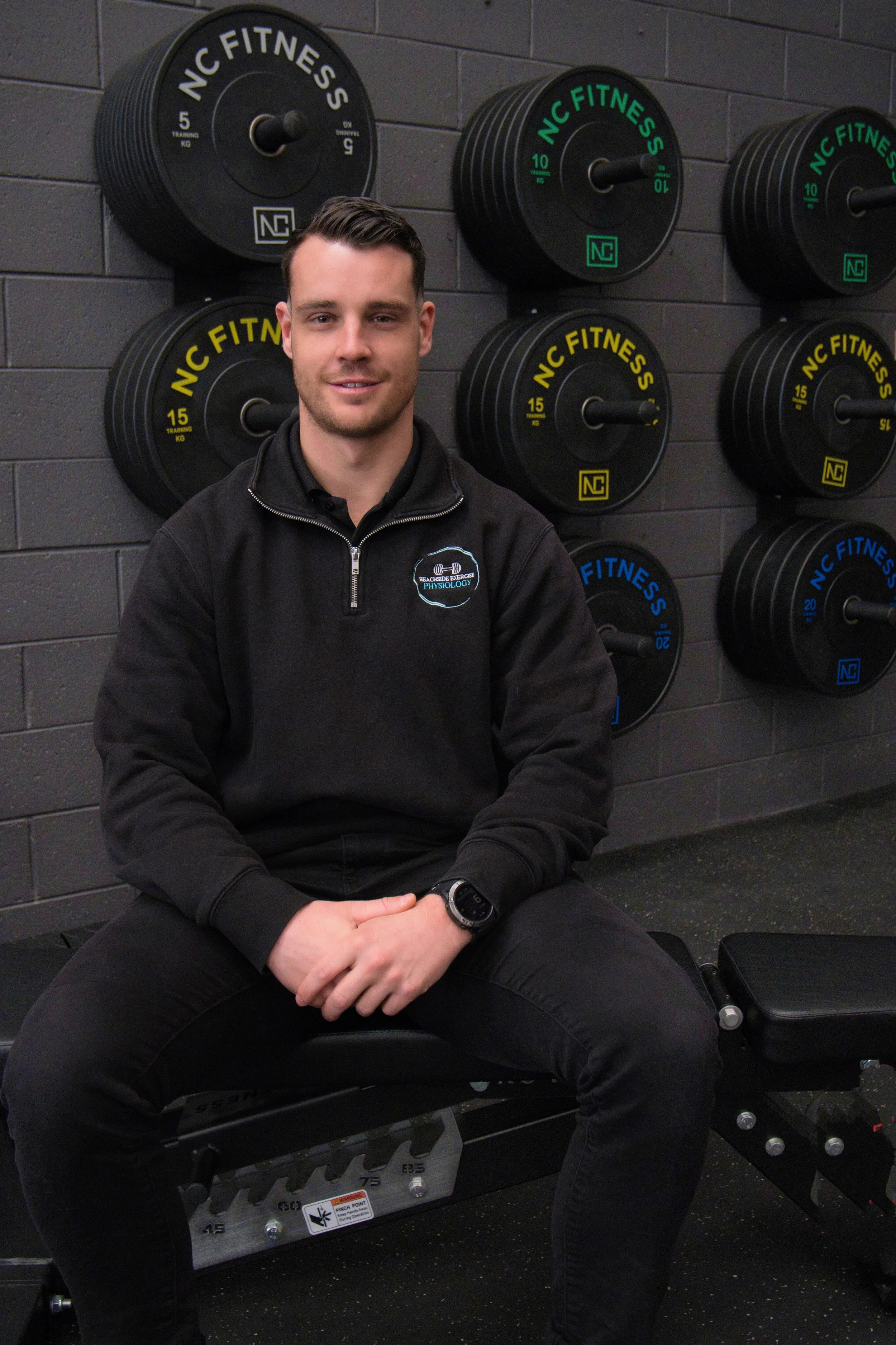 An accredited exercise physiologist sits on a bench in a gym, with Weightlifting plates labeled 'NC Fitness' mounted on a gray brick wall behind him. - Braeden King AEP