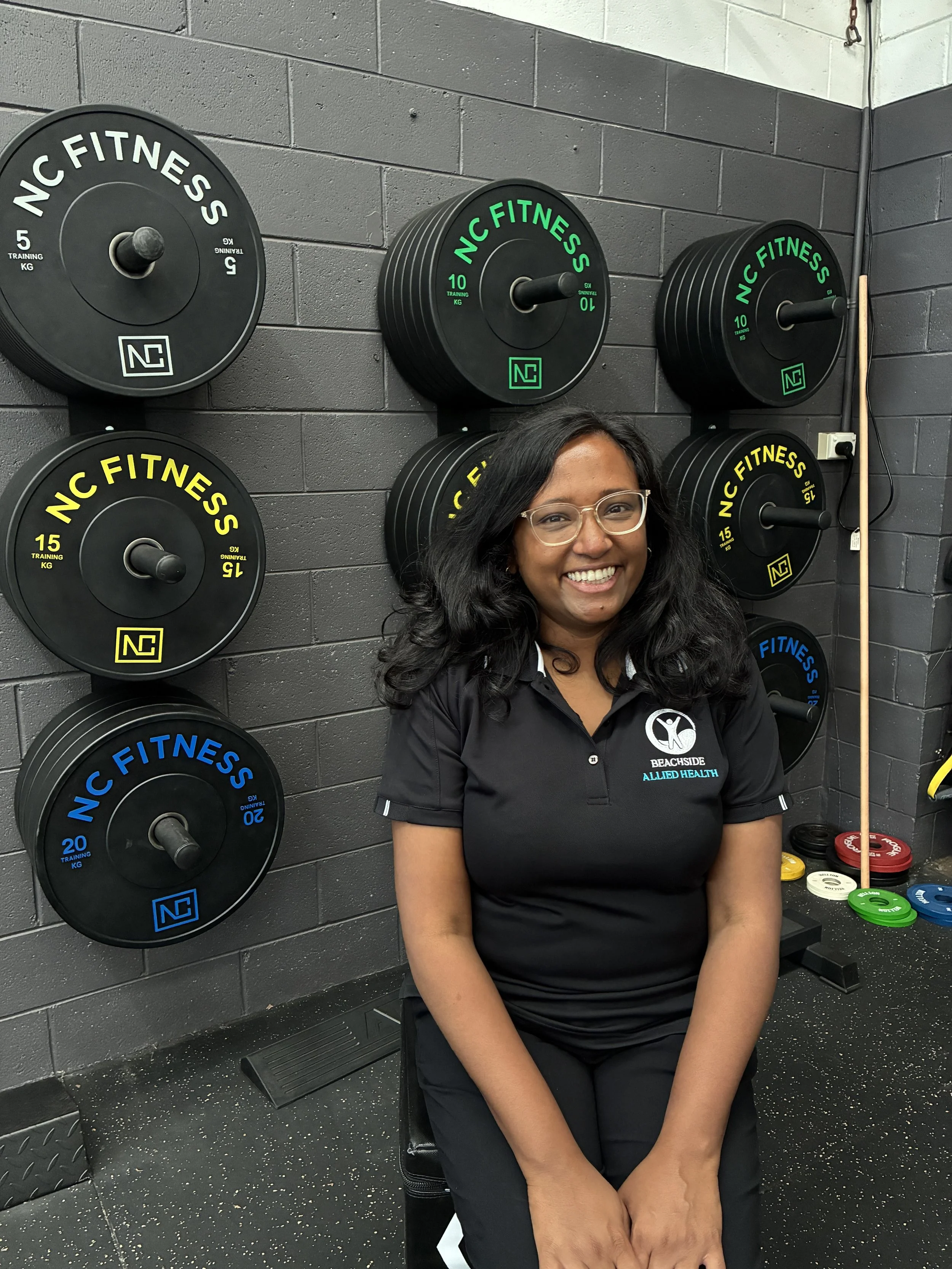 A woman sitting in a gym with a black shirt bearing the logos of Beachside Allied Health, smiling at the camera, in front of a wall with weight plates in black, green, yellow, and blue marked with weight measurements, and various colored weights on the floor.