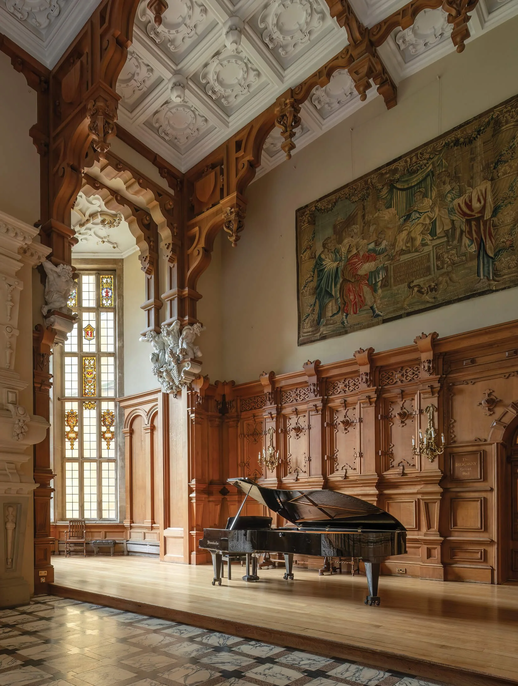A grand room with wooden paneling, a large window with stained glass, a black grand piano, a tapestry on the wall, and ornate decorations including sculptures and chandeliers.