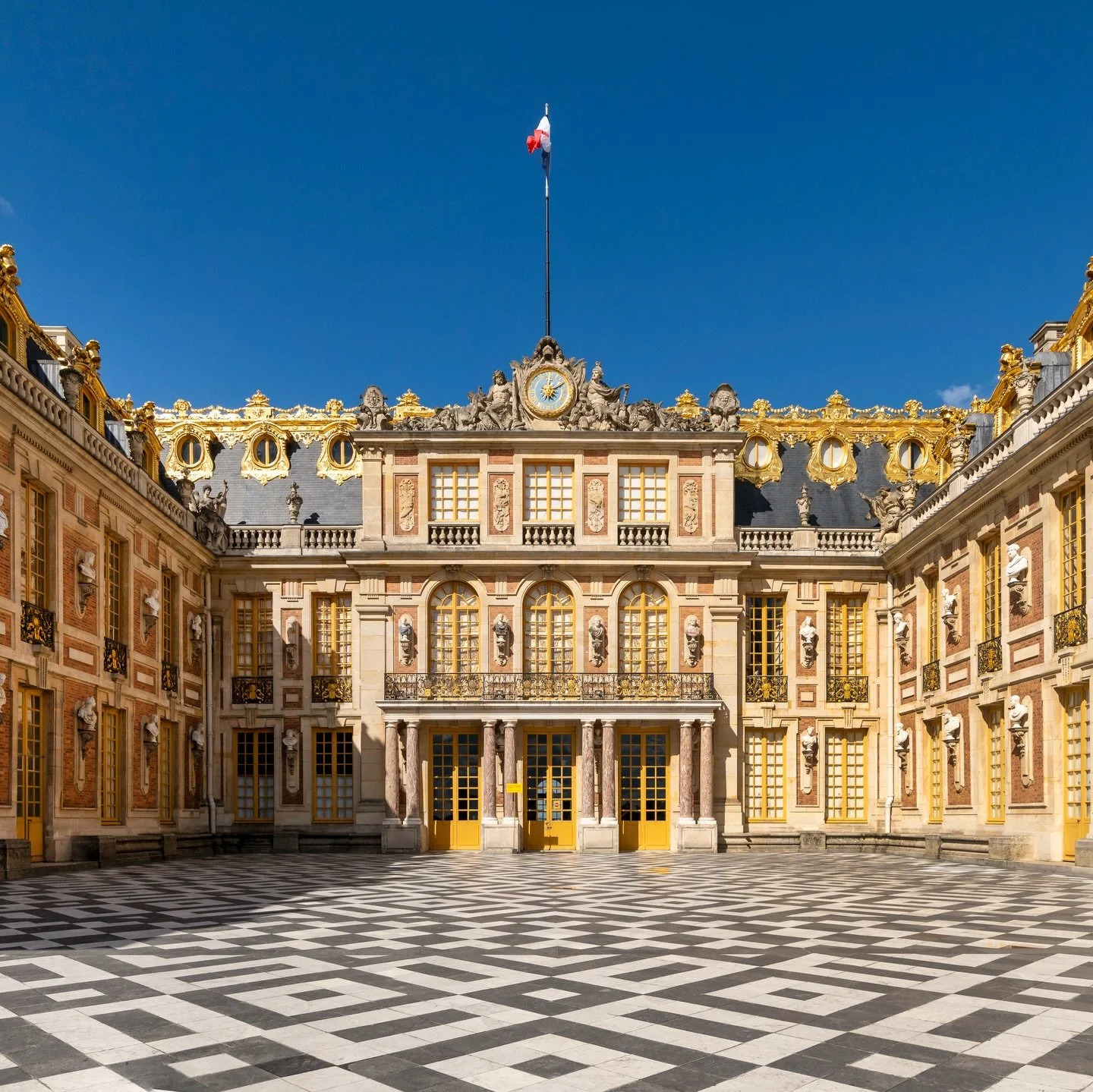 Photography of the exterior of the Palace of Versailles. The building is defined by its long classical fa&ccedil;ades of stone and brick, gilded ironwork gates, and a vast symmetrical courtyard designed to reflect the power and order of the French mo