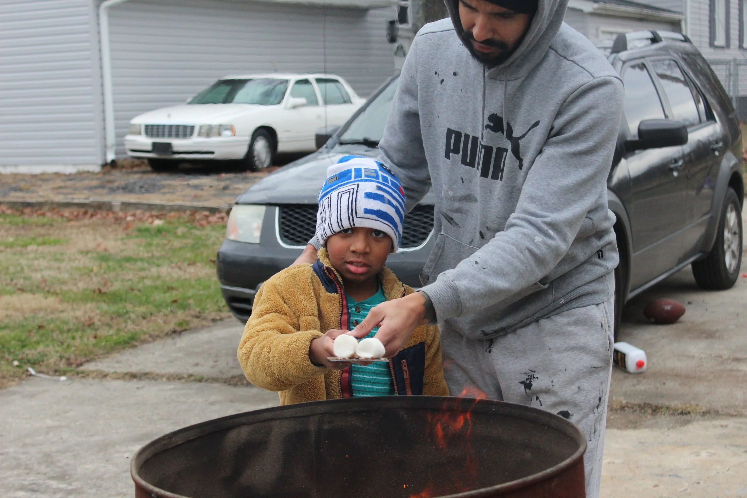 A man and a young boy standing next to a grill outdoors, with the man cooking and the boy holding marshmallows on a tray. The boy wears a striped knit cap and a tan jacket, and the man is in a gray hoodie and sweatpants, with cars parked in the backg