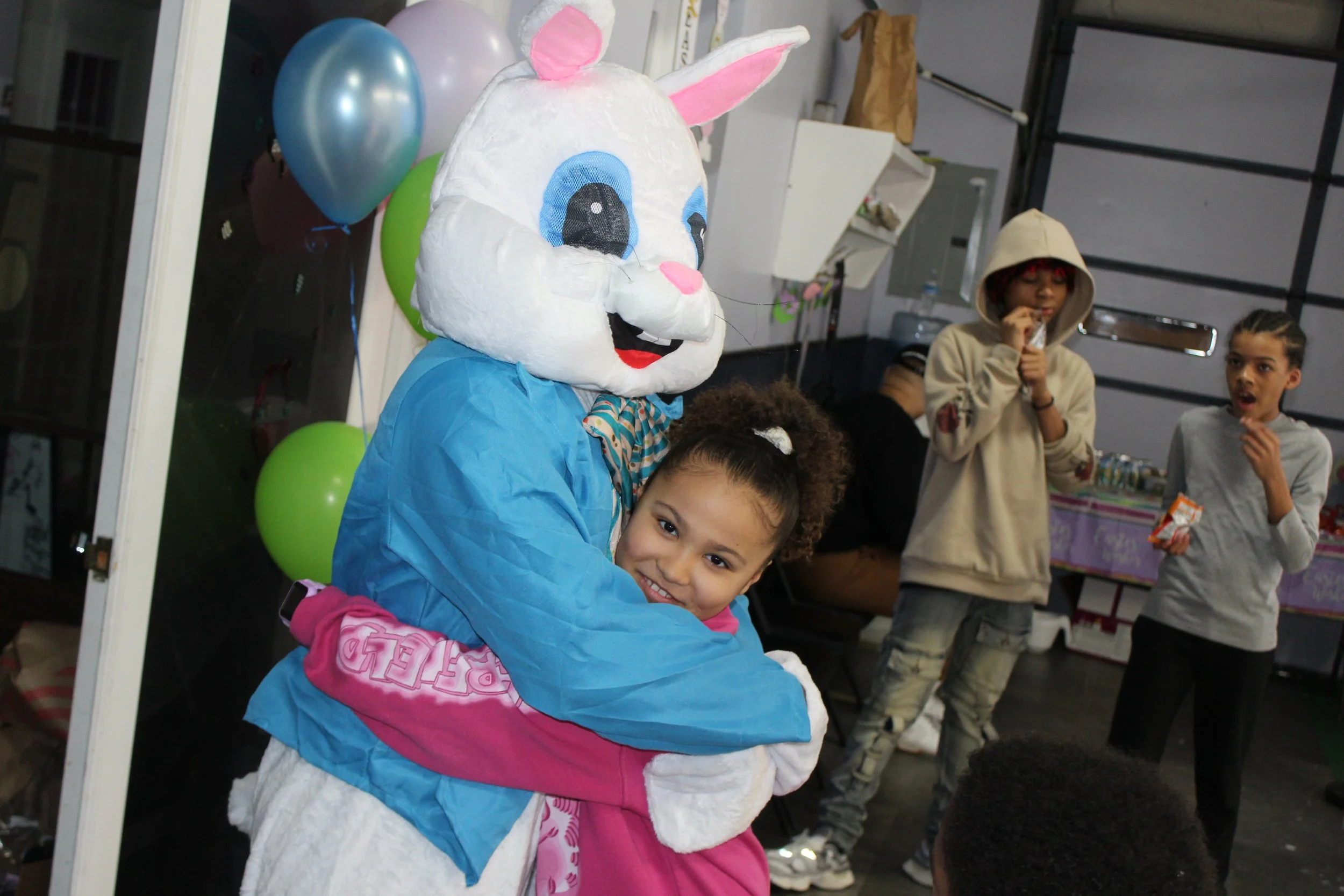 A young girl hugging a person in a bunny costume at a party, with balloons in the background and other children in casual clothes.