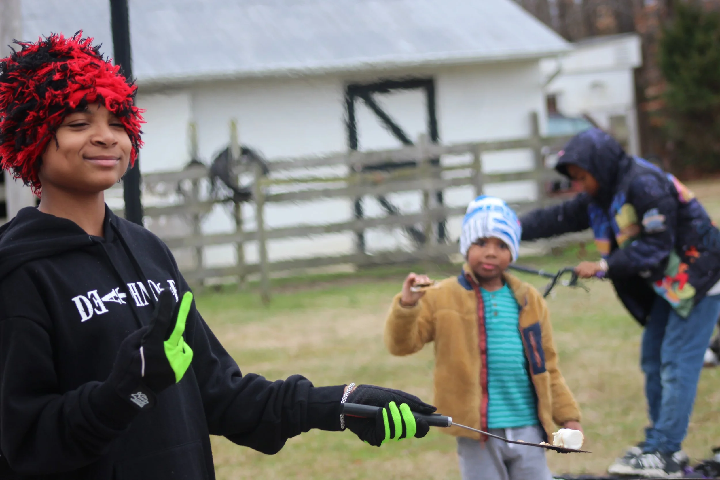 Three children outdoors, one in the foreground wearing a red and black fuzzy hat and black hoodie giving a thumbs up, the other two in the background with one holding a skewer with marshmallow and the third reaching to grab it, all dressed warmly, wi