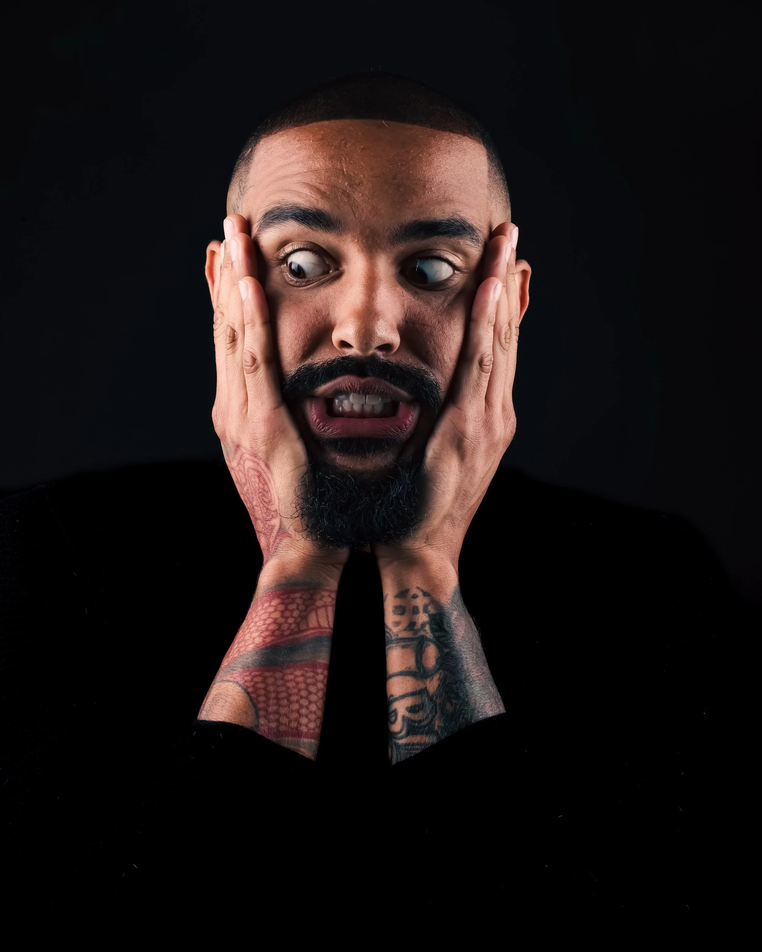Man with tattoos on arms holding his face with surprised or shocked expression against a black background.