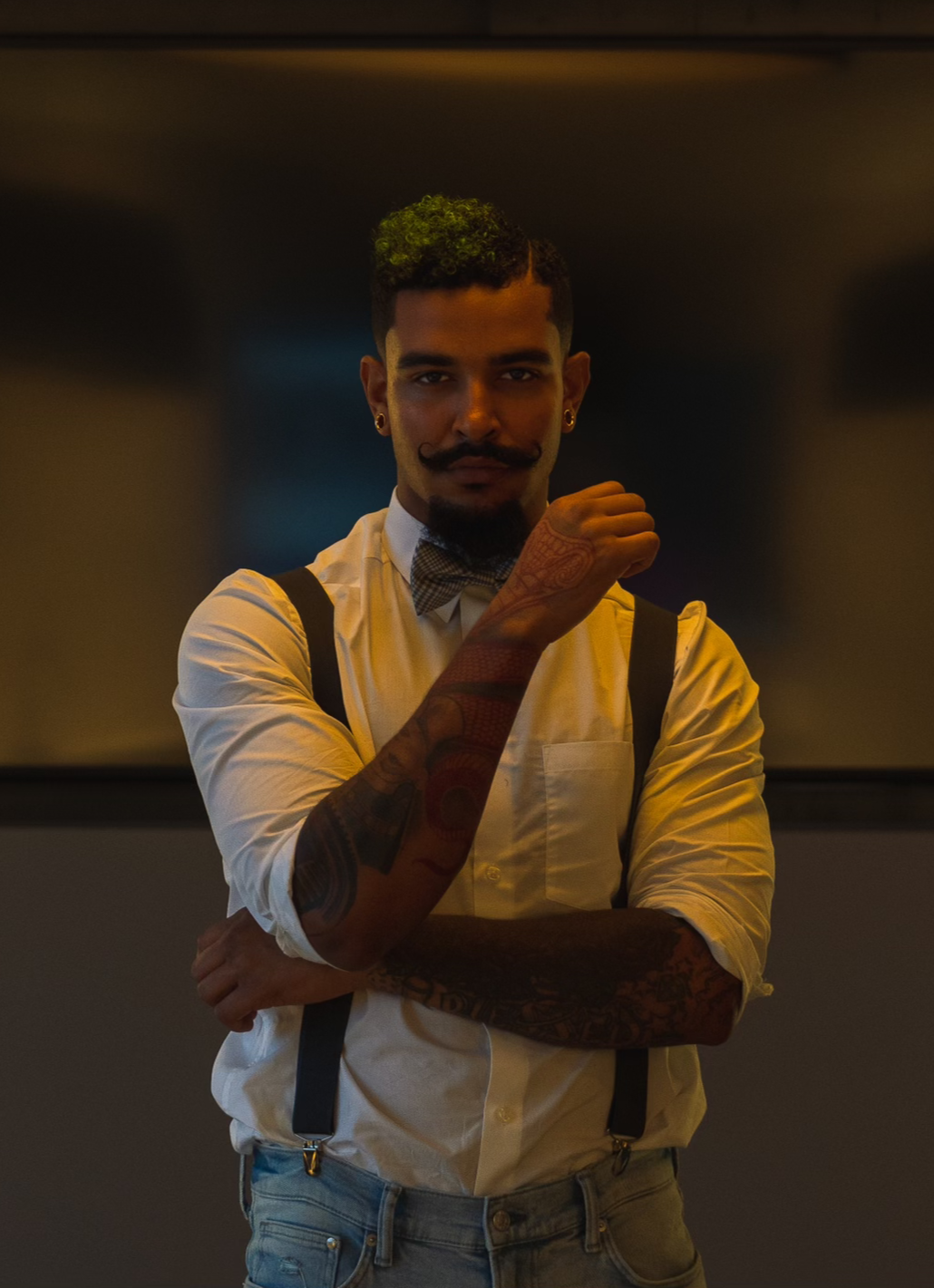 A man with styled hair, facial hair, and tattoos is standing with arms crossed, wearing a white shirt, suspenders, and a bow tie, in a dimly lit setting.