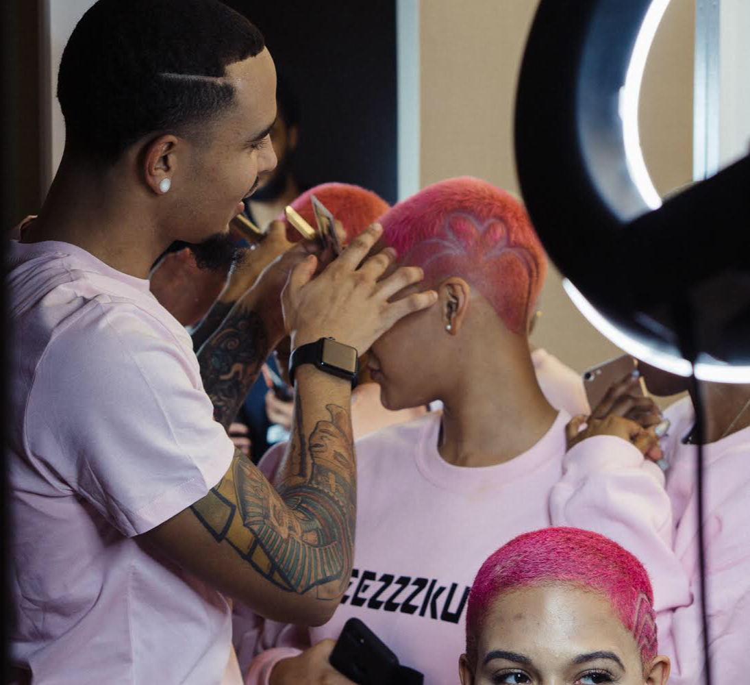 A woman with short pink hair getting her hair styled by a hairstylist with tattoos, in a salon or backstage area, with a large ring light illuminating the scene.