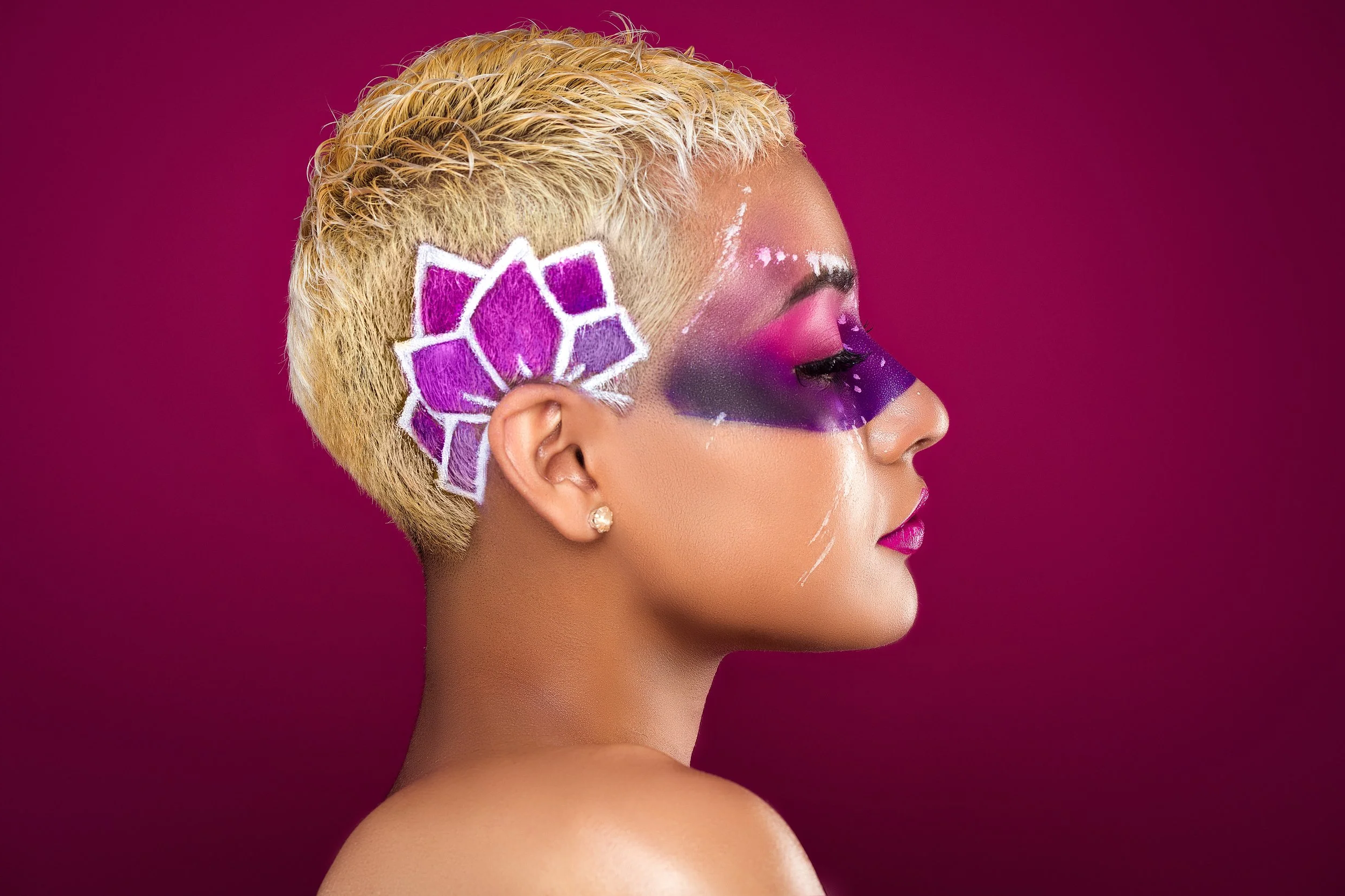 Side profile of a woman with short blonde hair, artistic purple and pink makeup, and a purple geometric design on the side of her head against a magenta background.