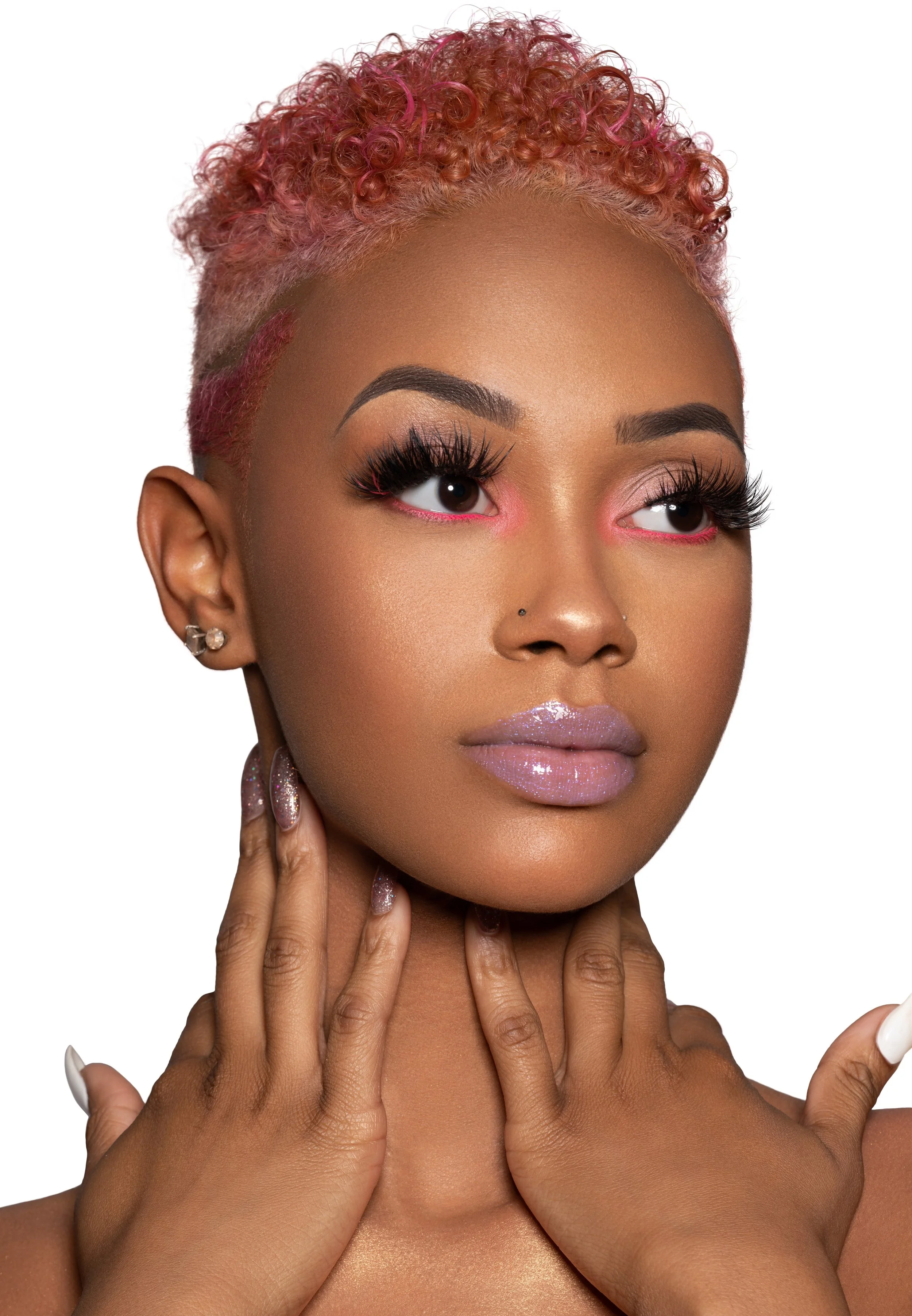 Close-up portrait of a woman with pink curly hair, glamorous makeup including pink eyeshadow, glossy lips, and long eyelashes, with her hands gently touching her neck.