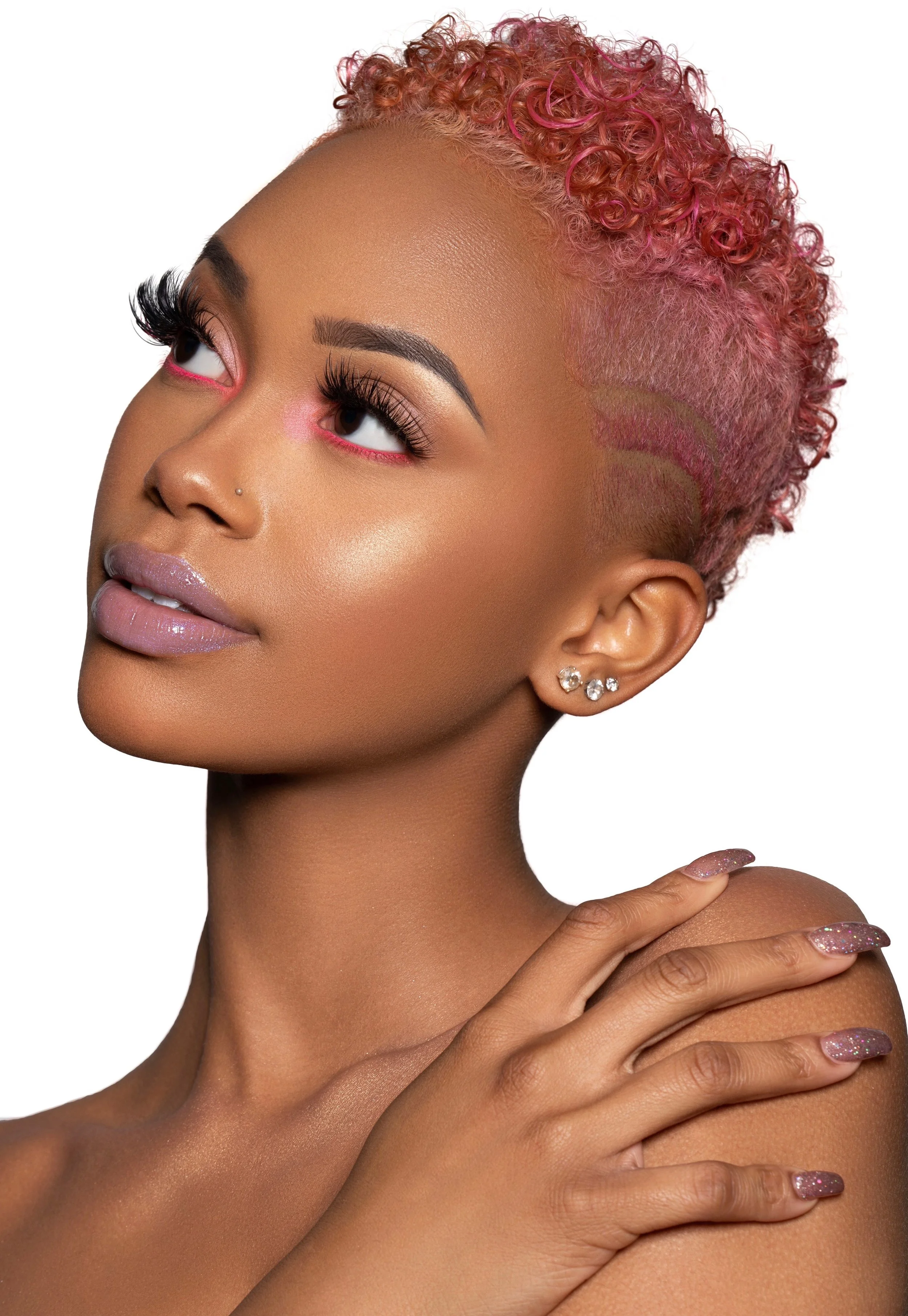 A woman with pink curly hair, makeup, and glittery nails, holding her shoulder with her hand, looking to the side against a white background.