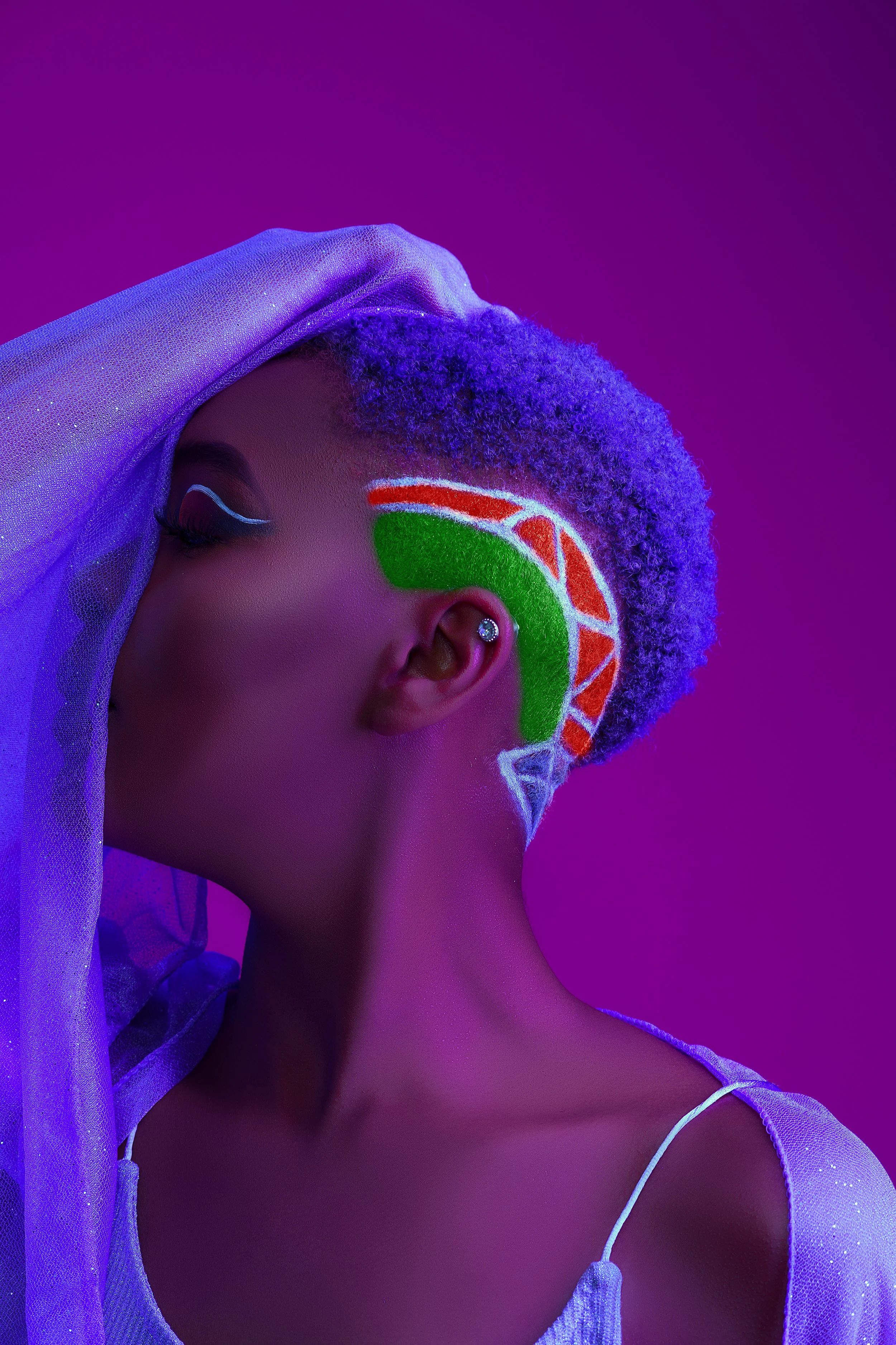 A person with dyed purple hair, vibrant face paint resembling a slice of watermelon, and a glittery purple sheer fabric draped over their head, against a purple background.