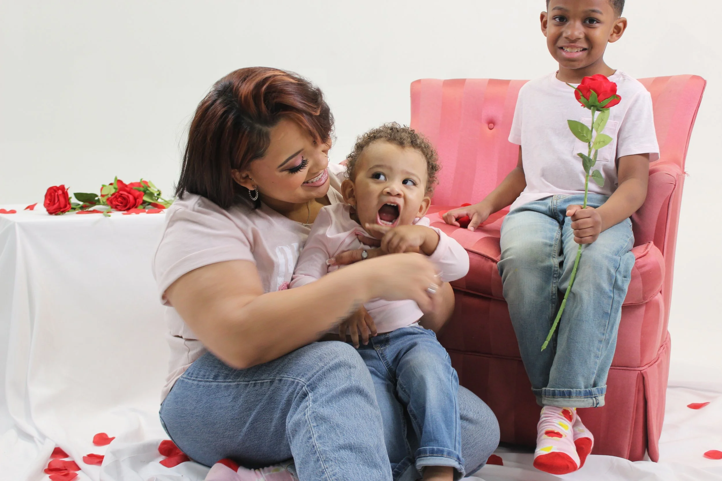 A woman playing with two young children on a pink couch, one child is smiling and holding a rose, and the other is making a funny face with her mouth wide open.
