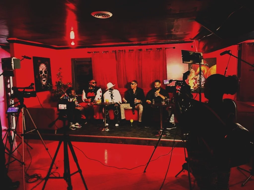 Group of six people seated on a stage with red walls, being filmed by multiple cameras, with artwork and curtains in the background.