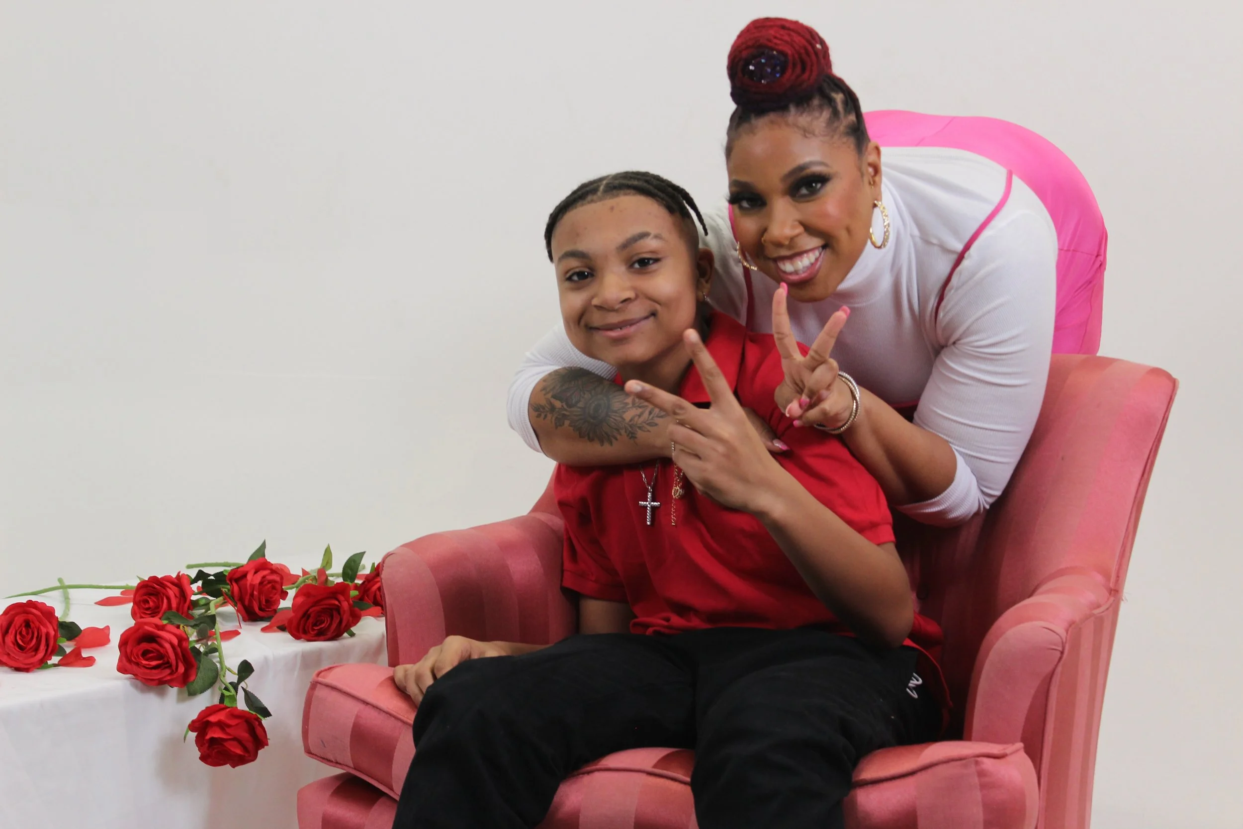 A woman and a young boy sitting on a pink armchair, making peace signs with their hands, with red roses on a table nearby