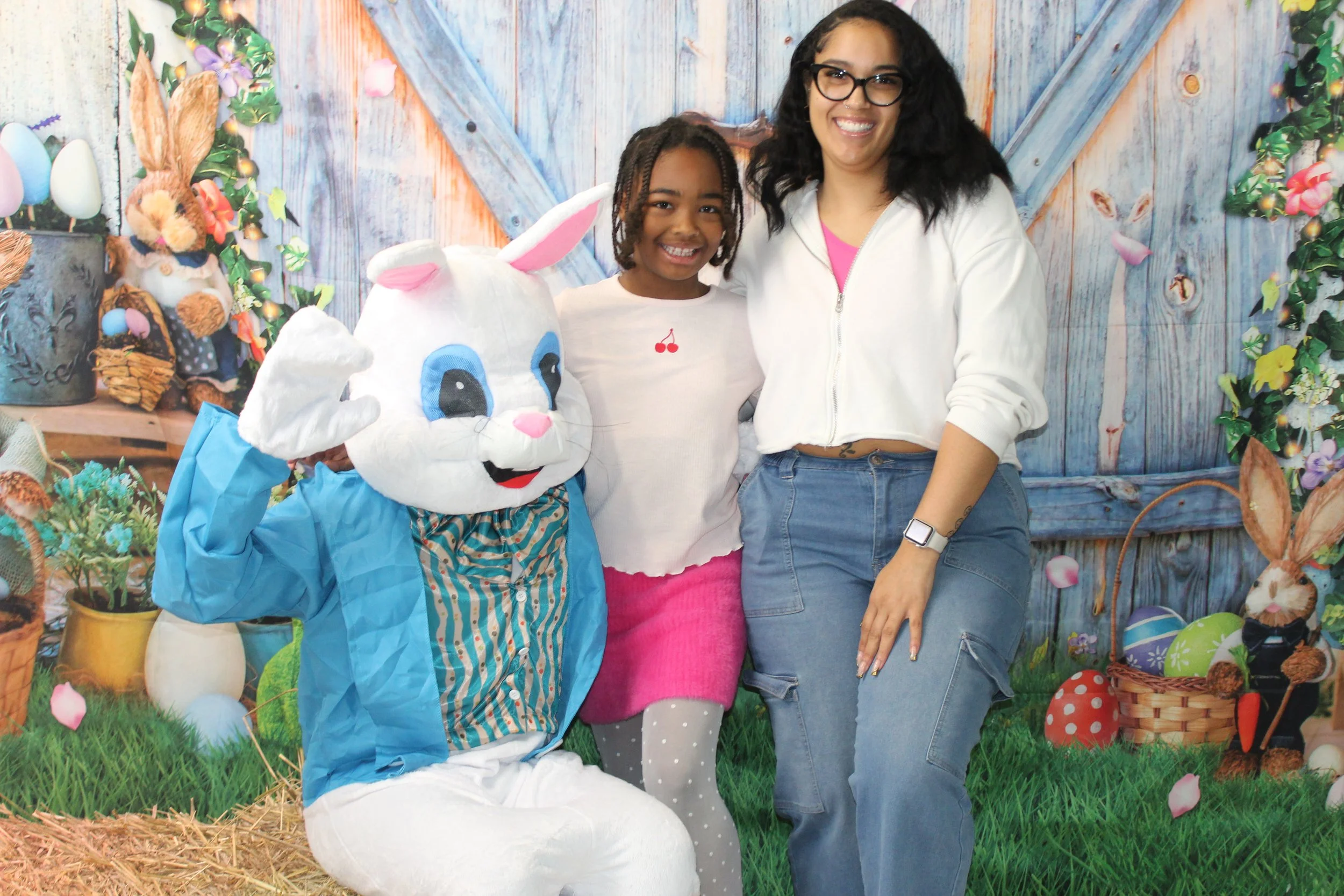 Three people, including a person in a bunny costume, standing in front of a decorated Easter-themed background with eggs, flowers, and grass.