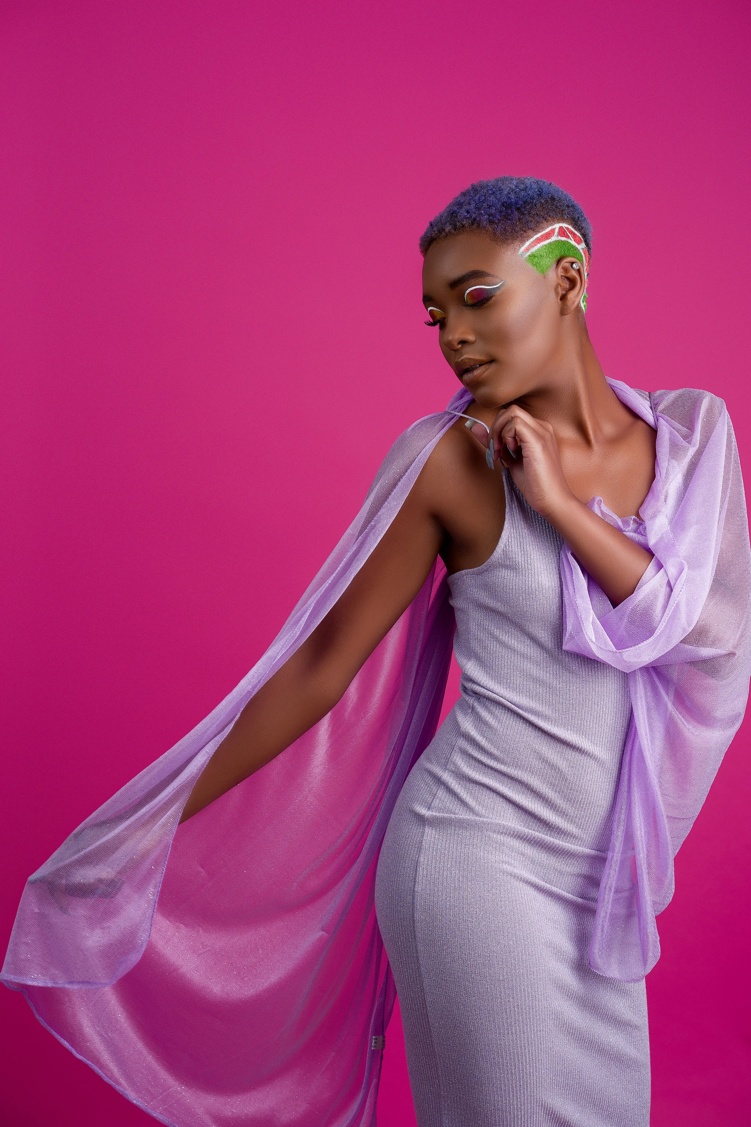 A woman with short, purple and blue hair, and colorful makeup, wearing a shiny, light purple dress and a sheer purple cape, standing against a pink background.