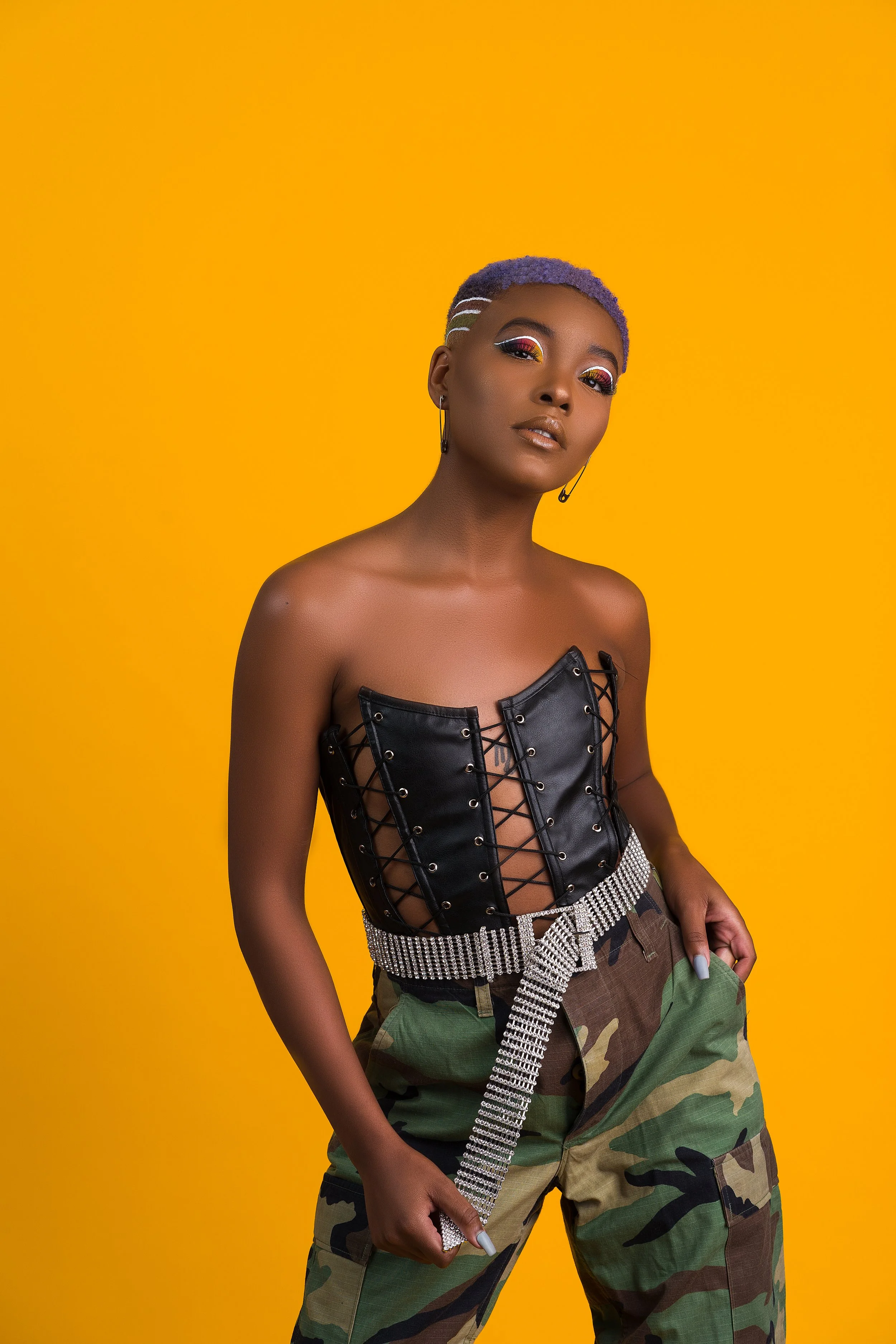 A woman with purple hair and colorful makeup poses in front of a yellow background, wearing a black leather corset with laces, camouflage pants, and a rhinestone belt.