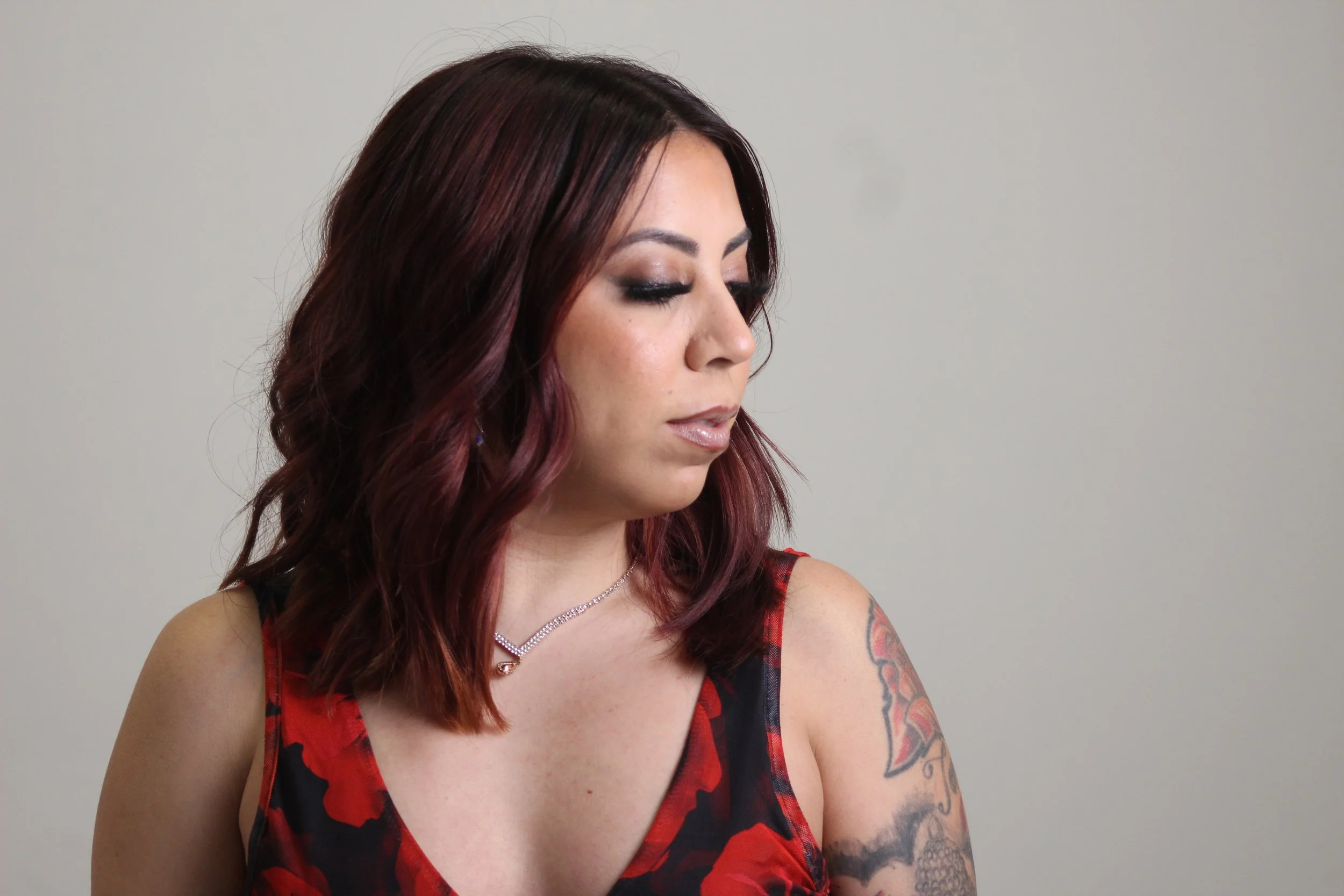 A woman with shoulder-length, wavy, burgundy hair looking down and to the side, wearing a red and black floral dress, a silver necklace, and a tattoo on her left arm, against a plain light gray background.