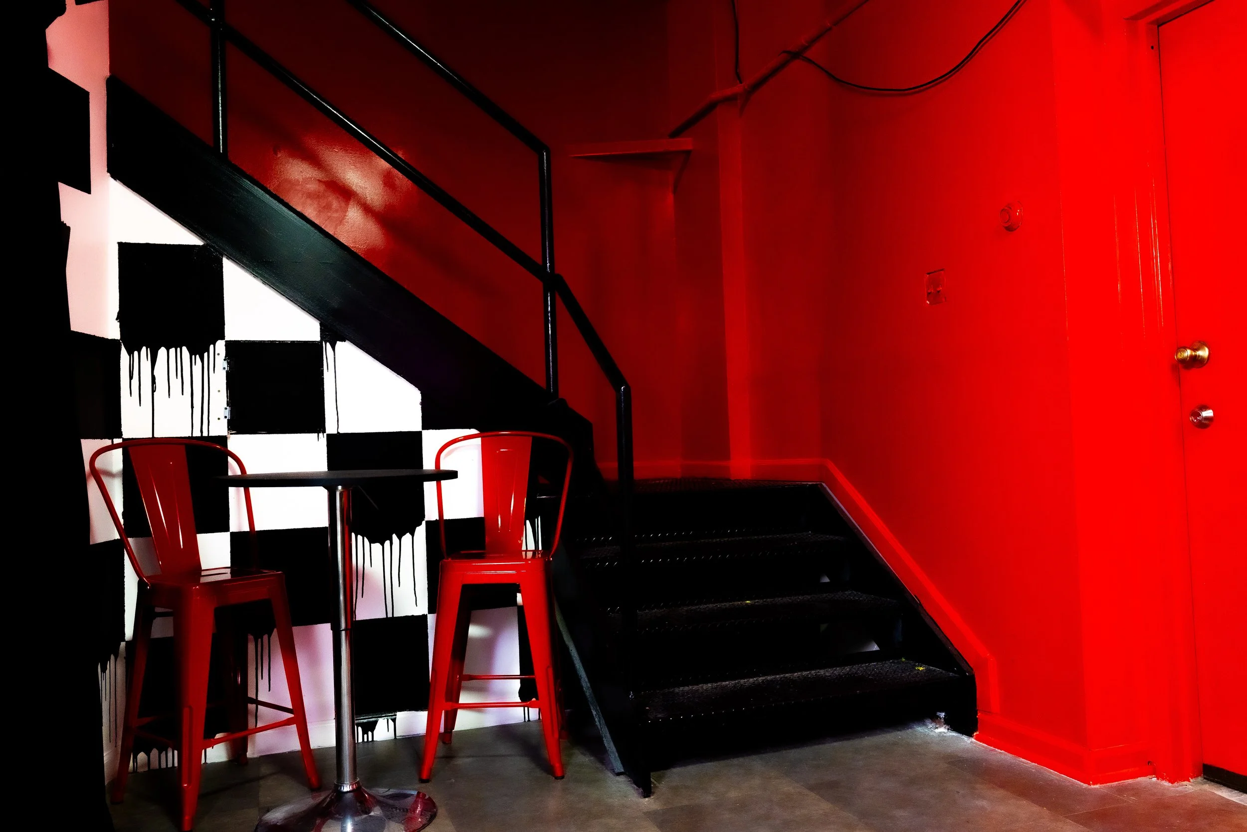 A small round table with two red chairs, black and white checkered wall with black drips, black staircase with a railing, and a red wall with a door in an indoor setting.