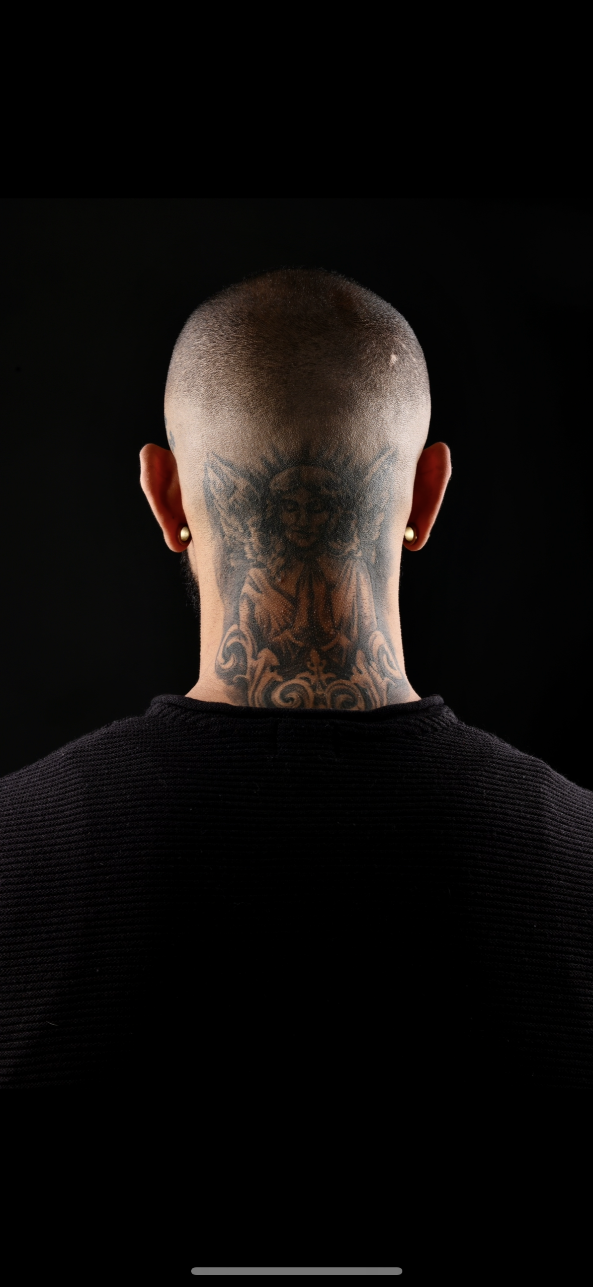 Back view of a person with a shaved head, tattoos on neck, wearing gold earrings and a black shirt, against a black background.