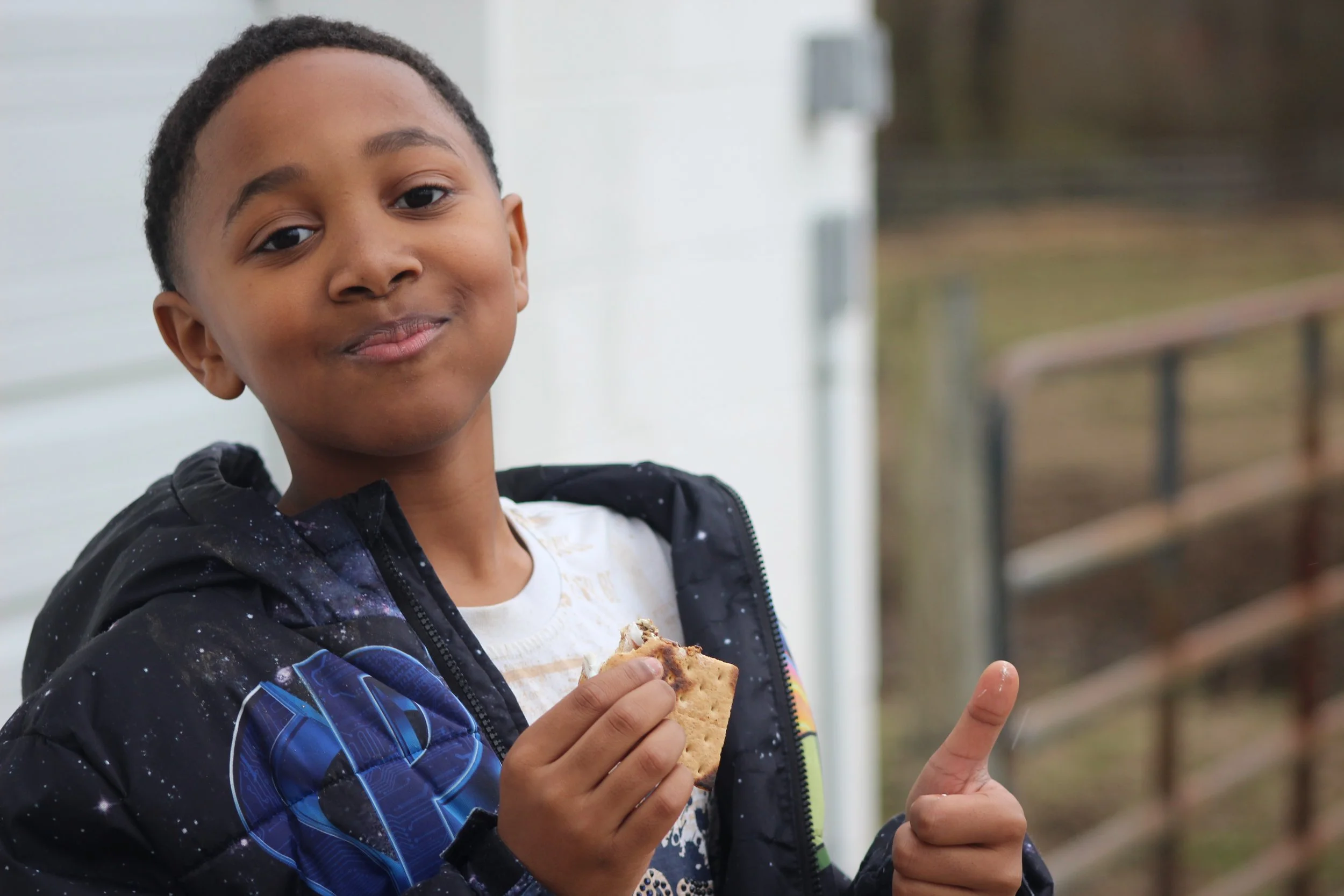 A young boy with short hair holding a cookie in one hand and giving a thumbs-up with the other, standing outdoors near a white wall with a blurred background of trees and a wooden fence.