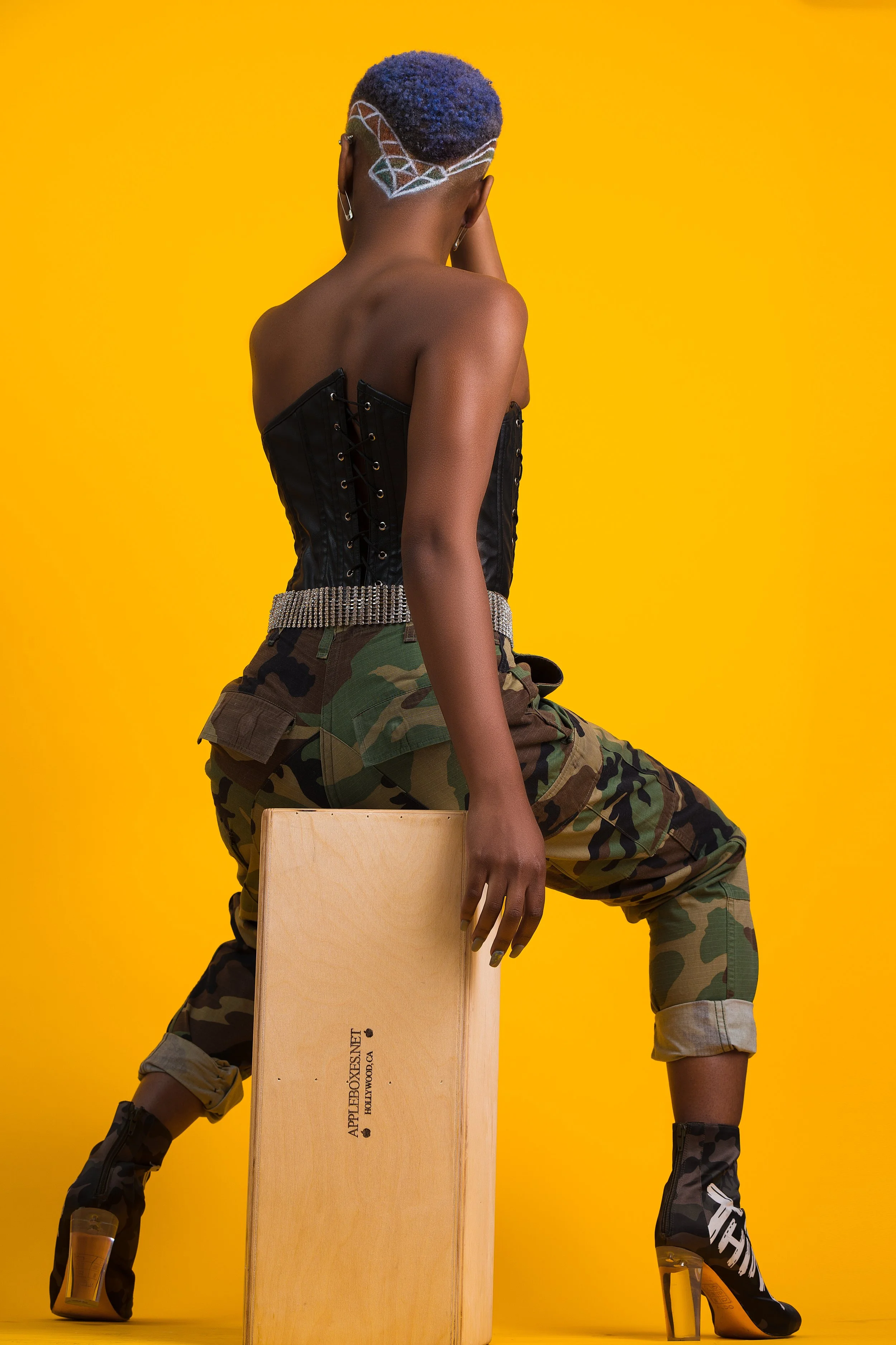 A woman with short, curly purple hair with a colorful geometric design shaved on the side, wearing a black corset, camouflage pants, and high-heeled combat boots, sitting on a wooden box against a yellow background.
