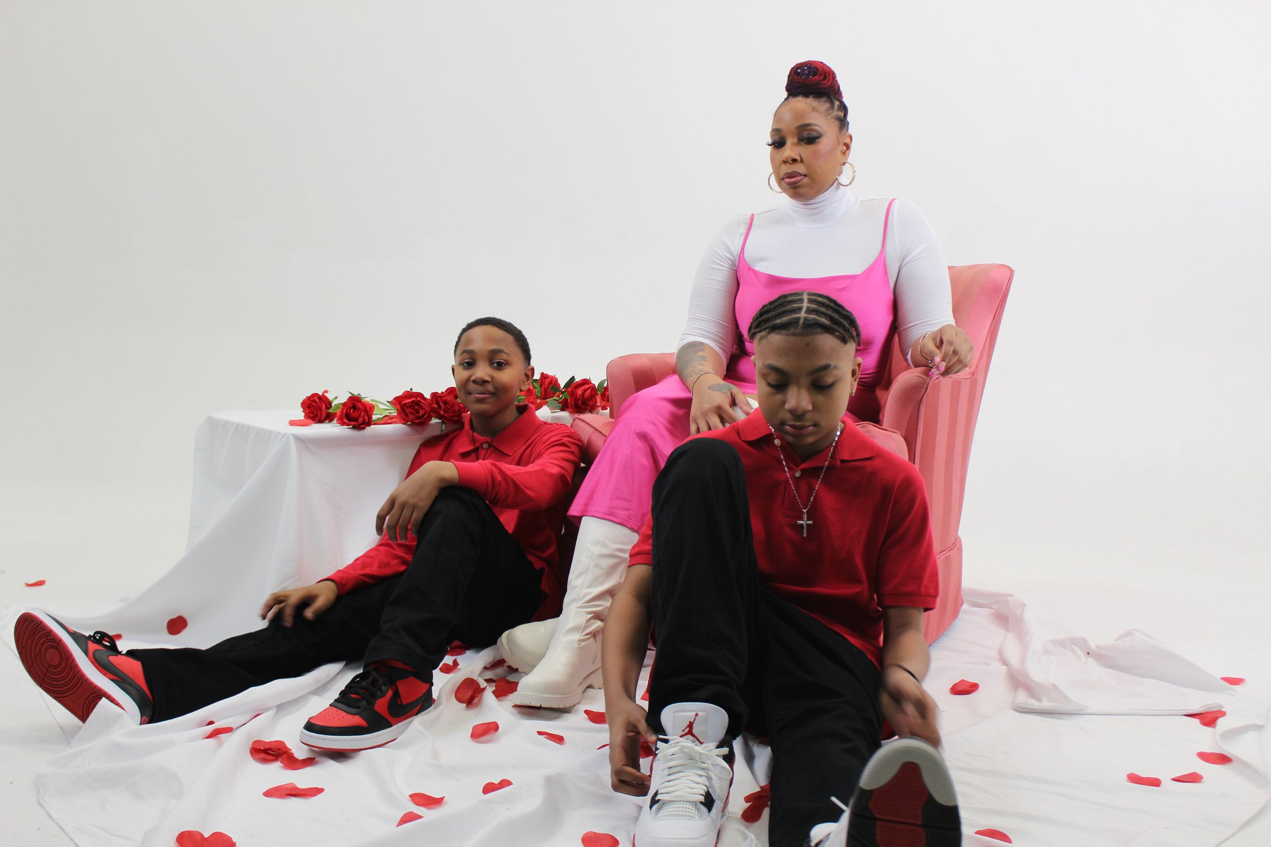 A woman with braided hair and hoop earrings sitting on a pink chair with two boys sitting on the floor in front of her. The boys are holding their shoes and the woman appears to be adjusting her shoe. There are scattered rose petals on the floor and 