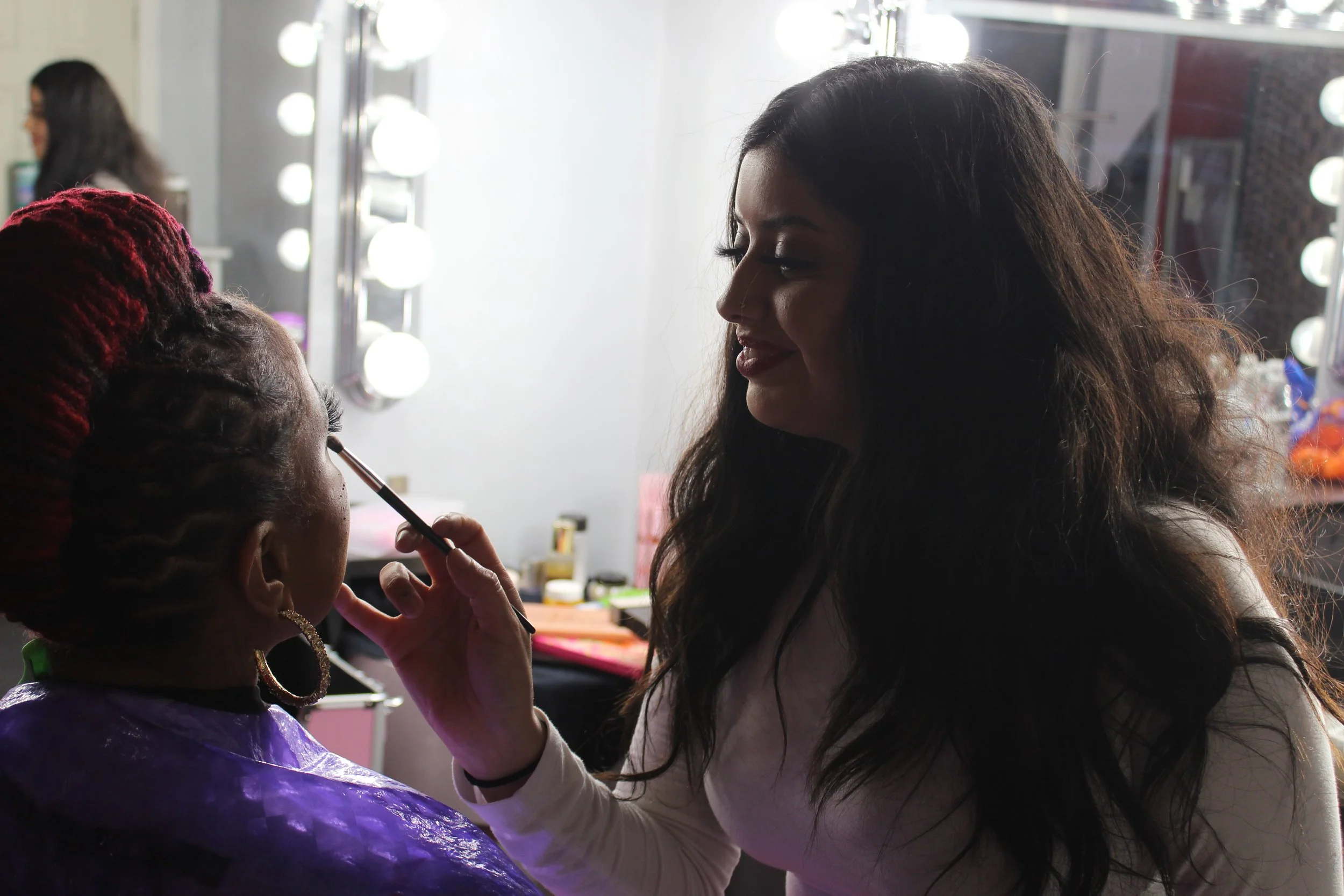 Makeup artist applying makeup to a woman in a dressing room with bright vanity lights.
