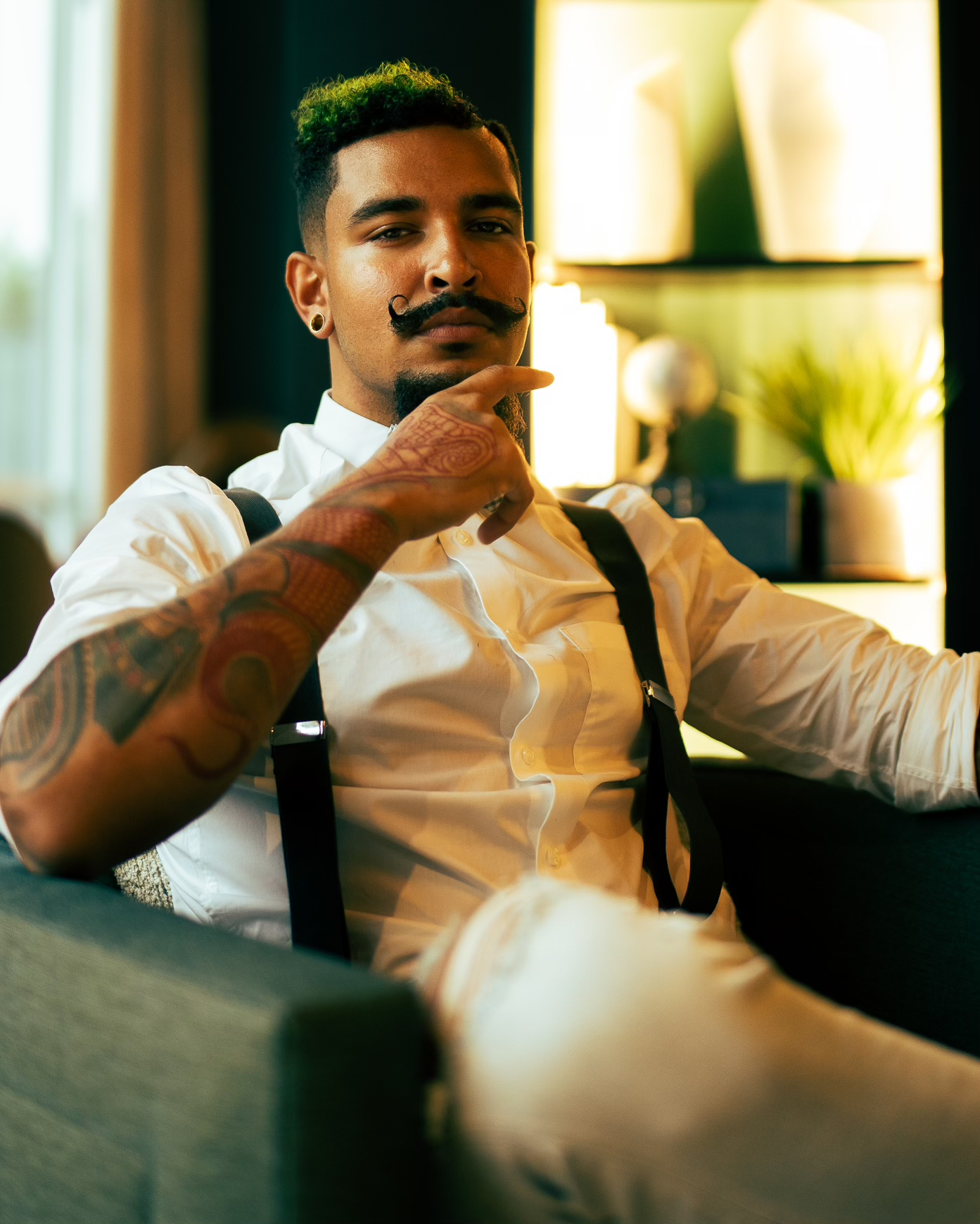 A stylish man with a mustache and tattoos, wearing a white shirt and suspenders, sitting on a sofa in a modern, well-lit room with plants and decor in the background.