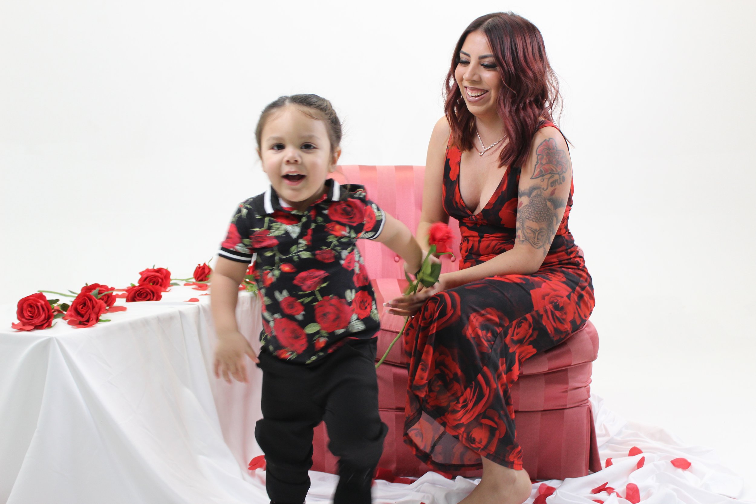 A woman with red hair and tattoos sitting on a pink chair, holding a red rose, smiling at a young boy in a floral shirt, with rose petals scattered on the floor and a table decorated with roses in the background.