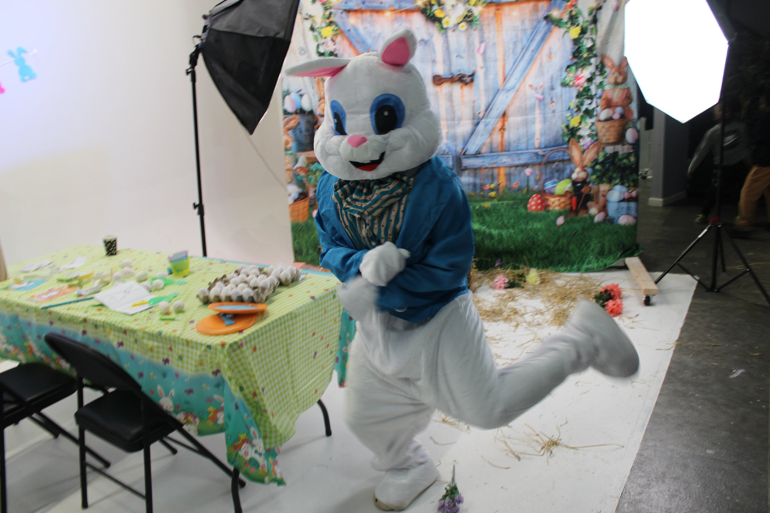 Person in a bunny costume with a large head, white fur, pink nose, and big blue eyes, dressed in a blue jacket and striped scarf, posing with one leg raised and crouched arms in front, at a decorated Easter-themed backdrop with a table of eggs and de