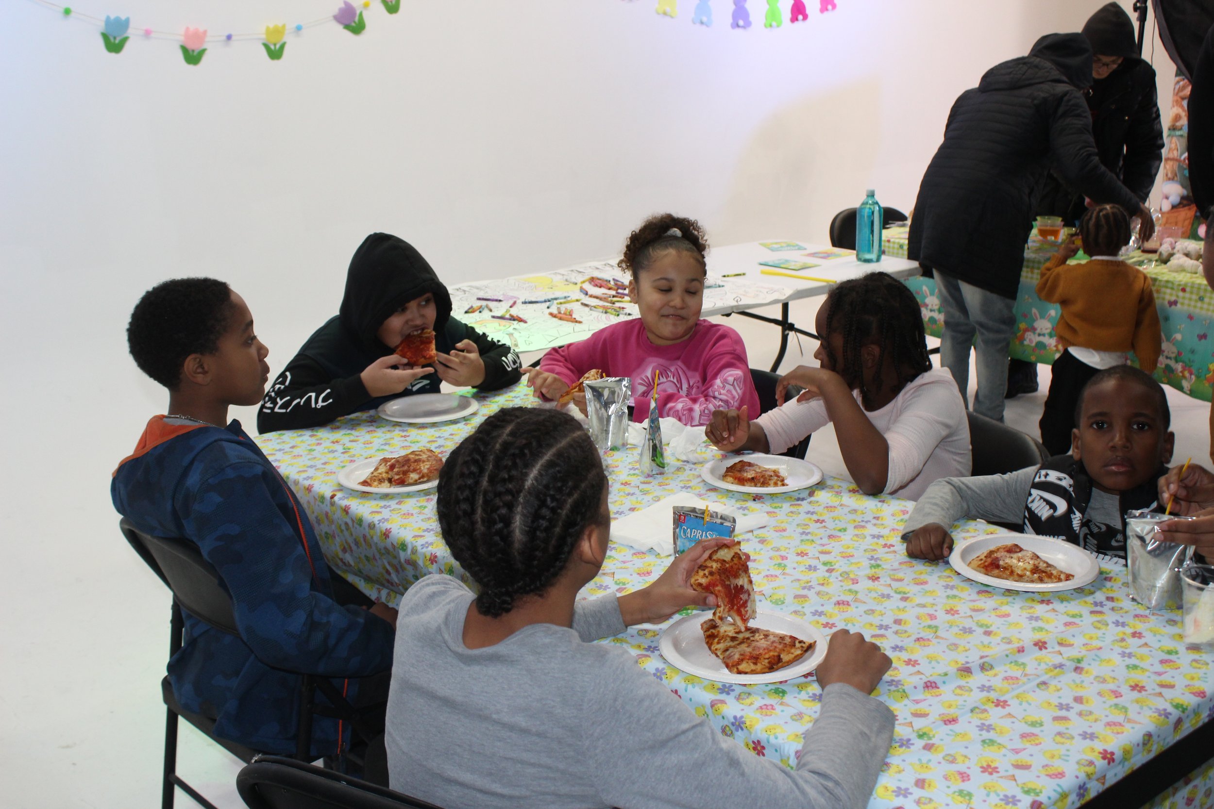 Children sitting around a table eating pizza at a party, with decorations and additional tables in the background.