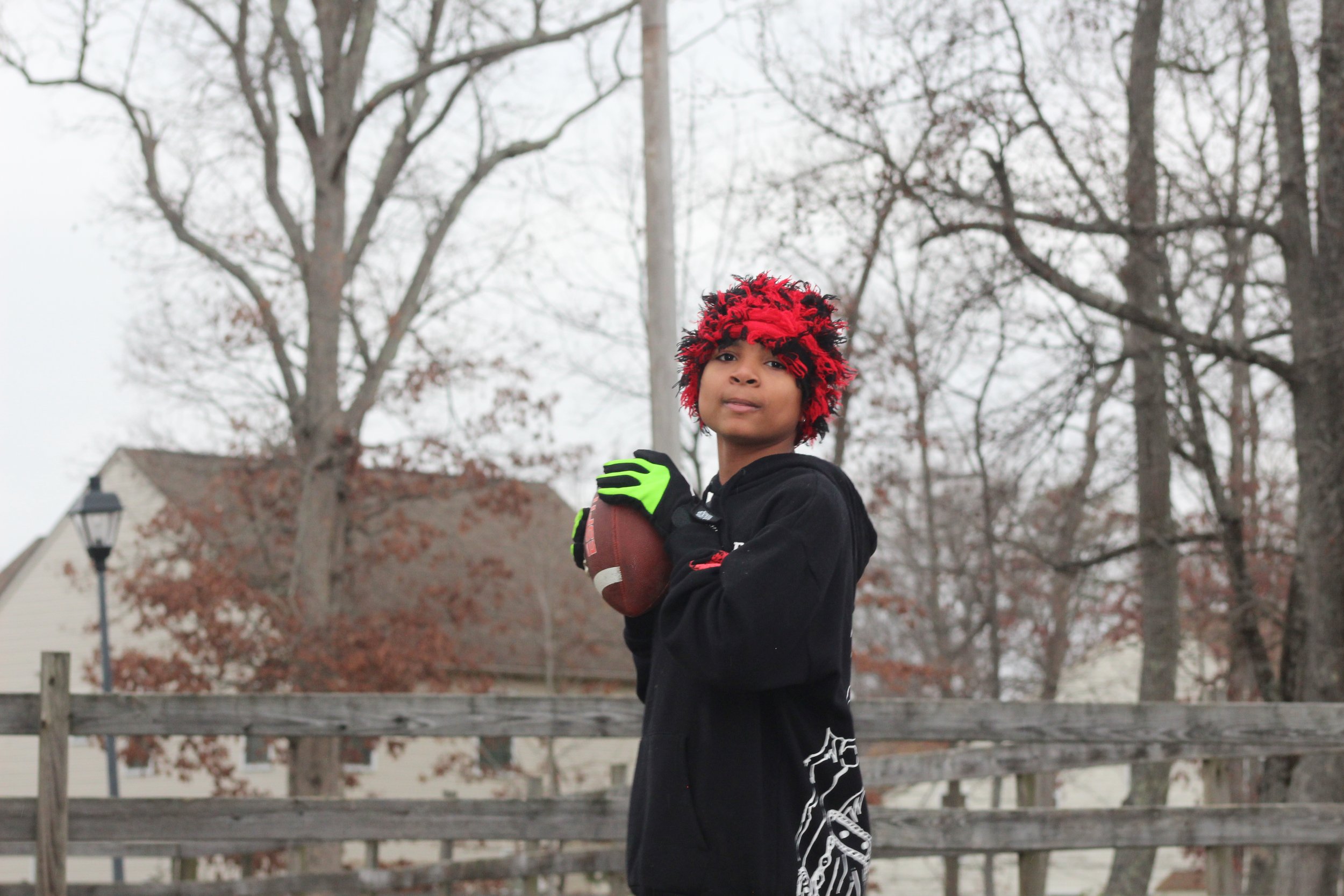 A young person with red and black curly hair wearing a black hoodie with graphics, neon green gloves, and holding a football, standing outdoors in a wooded area with leafless trees and a wooden fence.