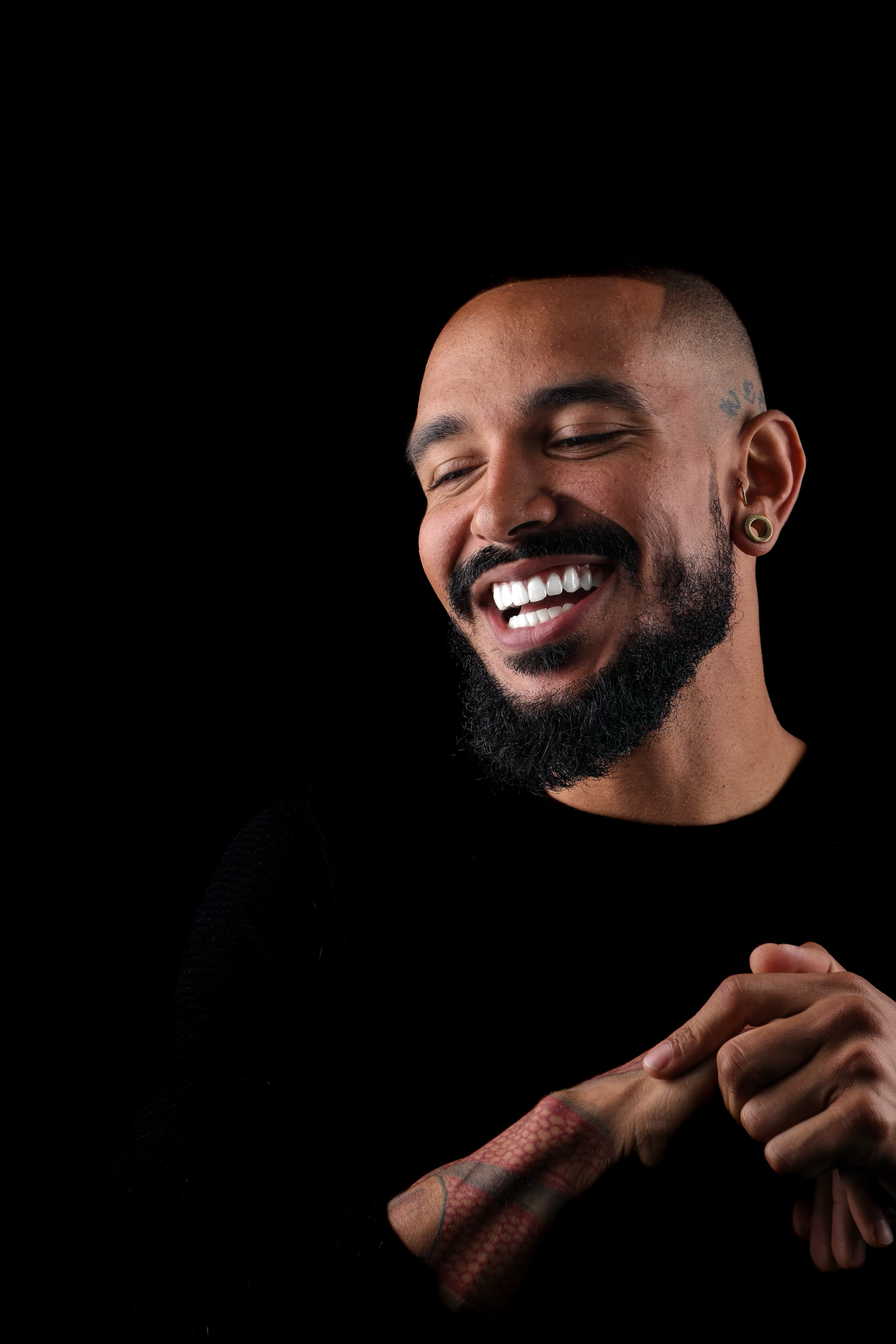 A man with a beard and tattoos smiling and looking down against a black background.