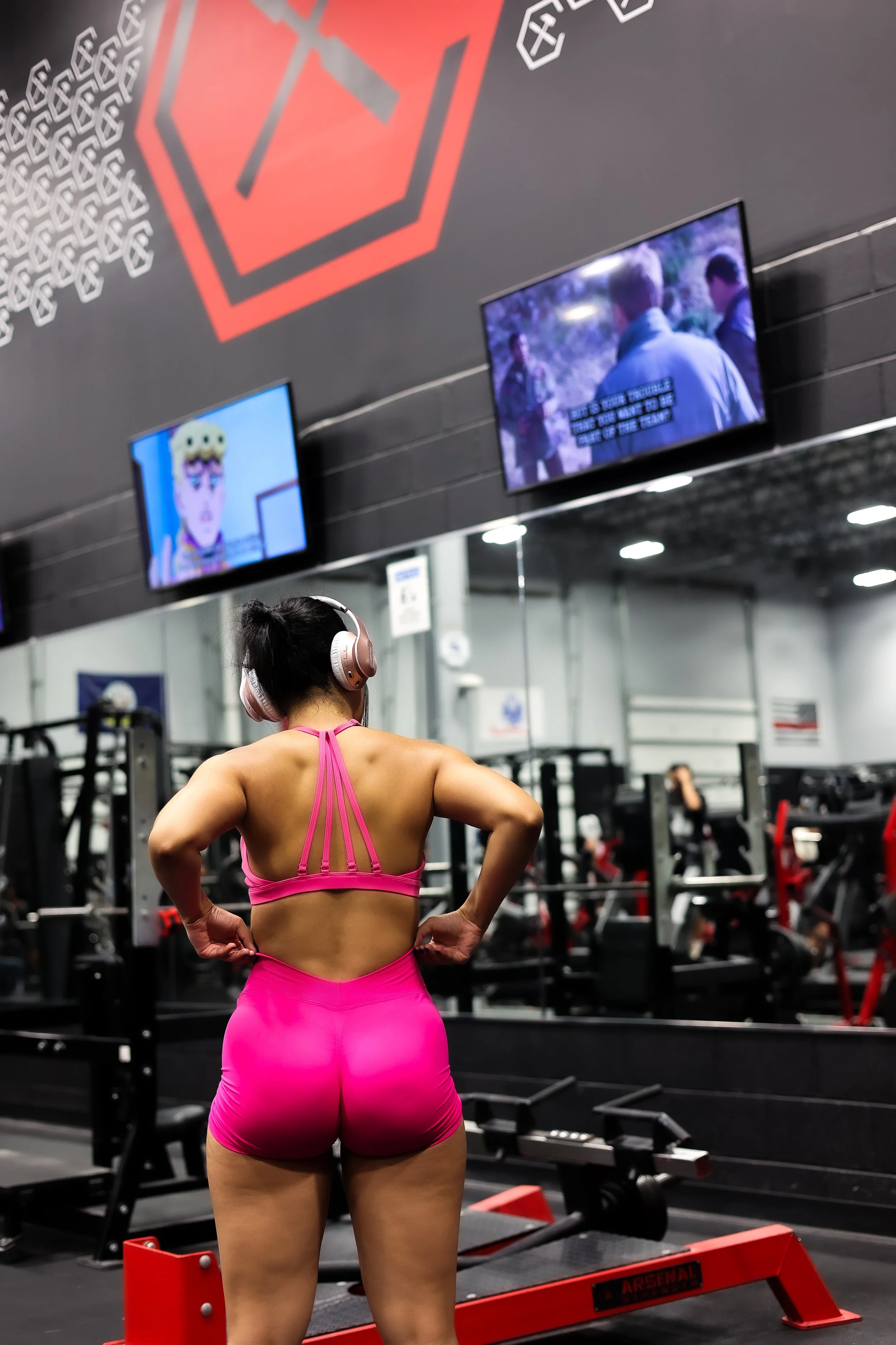 A woman in a pink workout outfit and headphones standing in a gym, facing mirrors and televisions, with her hands on her hips.